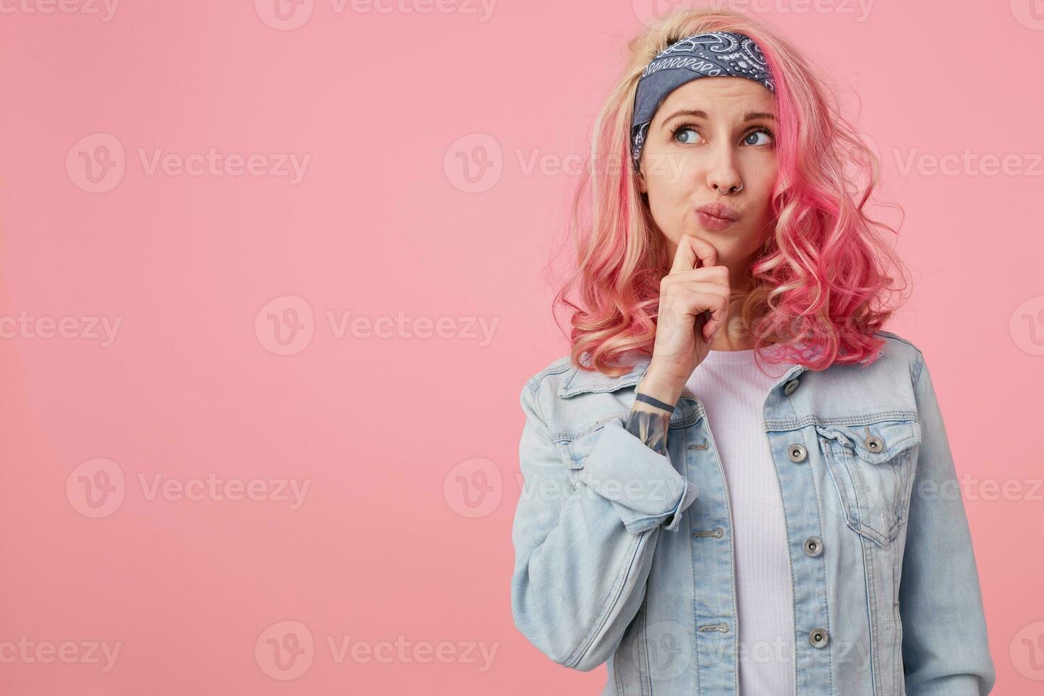 Young thinking cute lady with pink hair, wears a white t-shirt and denim jacket, stands over pink background copy space, looks up thoughtfully and touches the cheek with his finger. photo