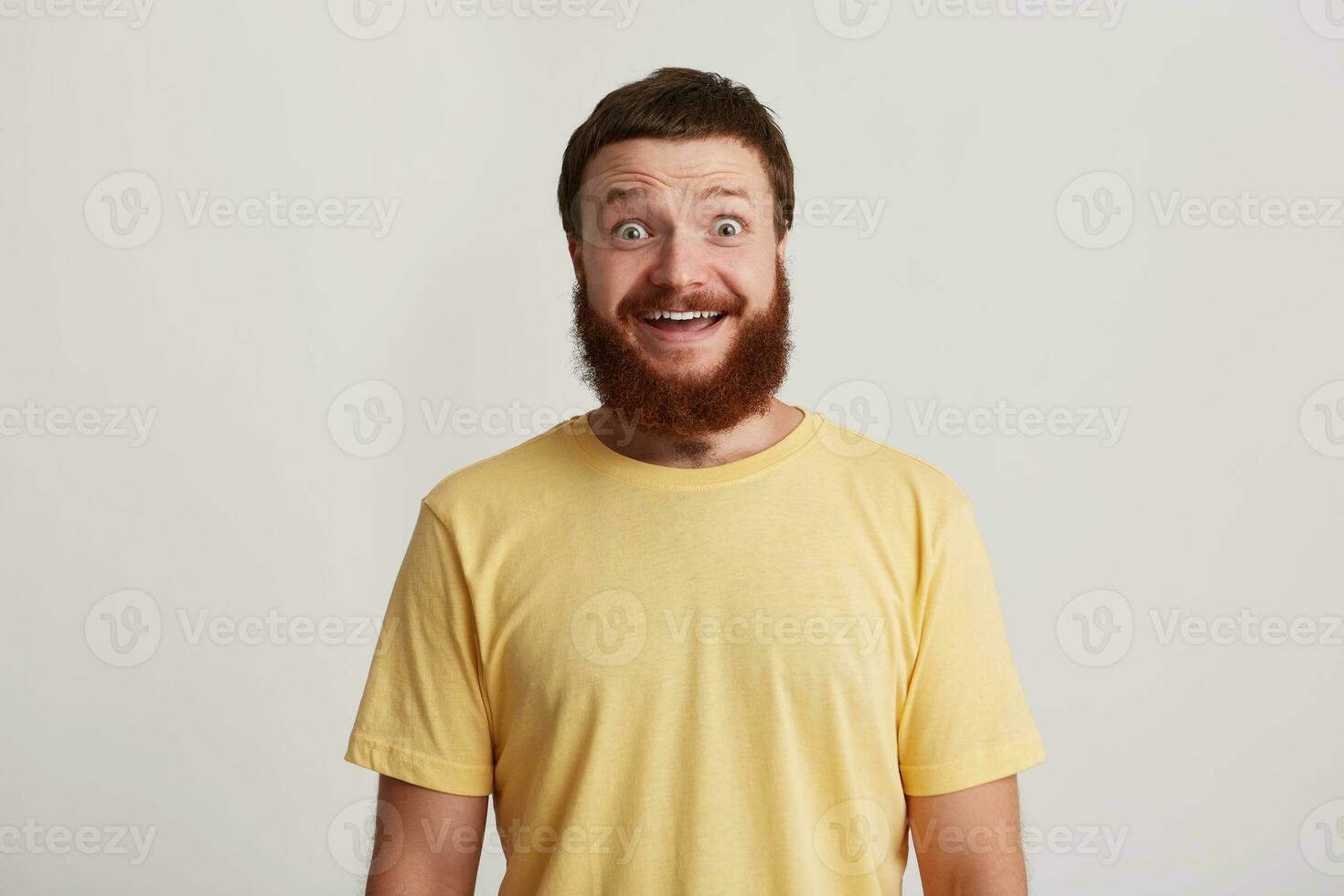 Portrait of happy surprised young man hipster with beard wears yellow t shirt standing with opened mouth and looks amazed isolated over white background photo
