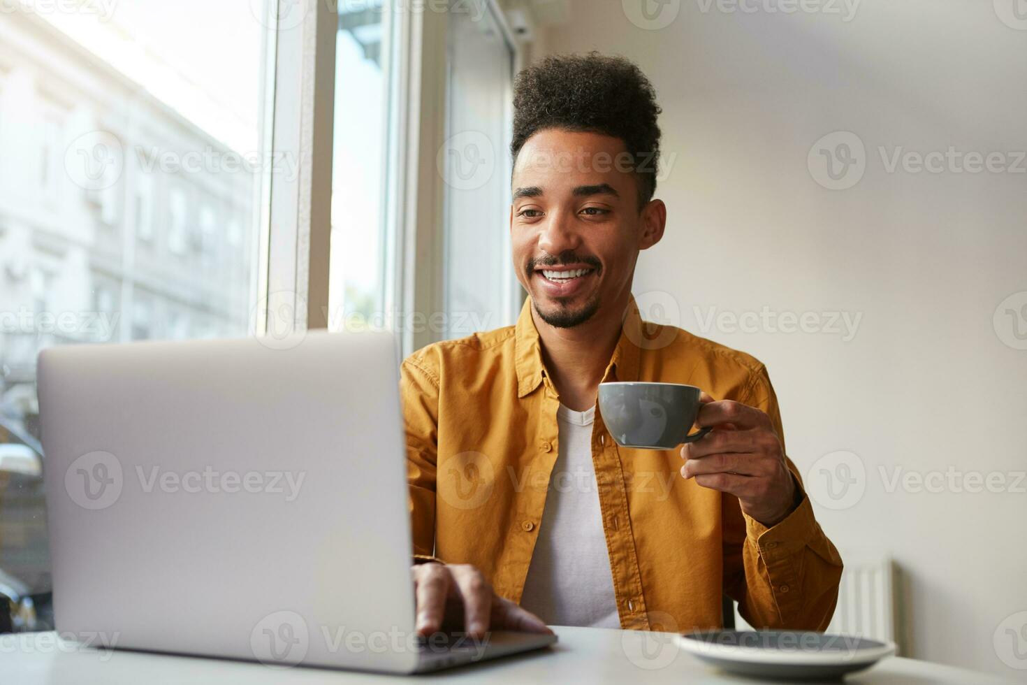 African American guy sitting at a table in a cafe and working on a laptop, wears in yellow shirt, drinks aromatic coffee, chatting with his girlfriend, enjoys the day. photo