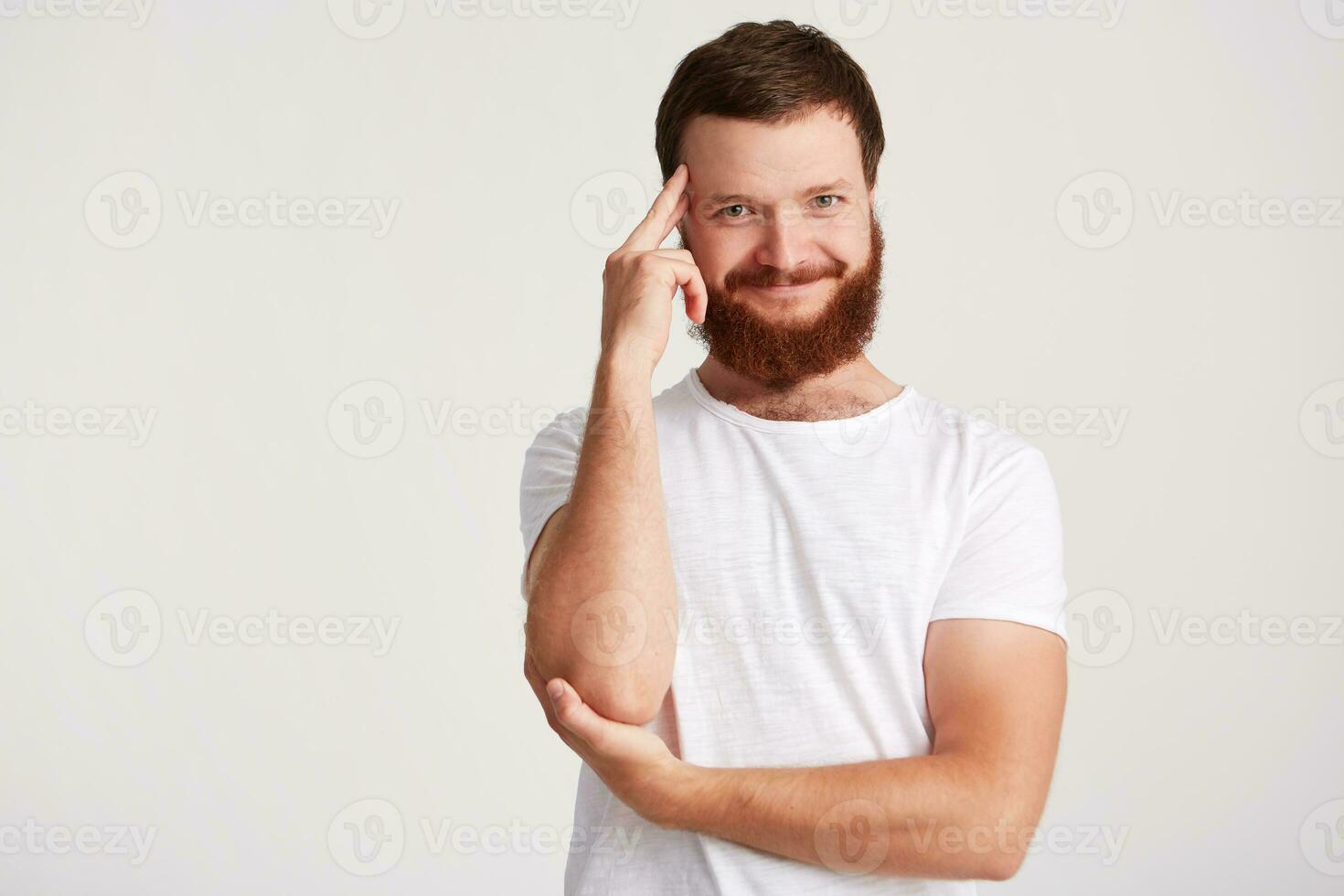 Portrait of smiling pensive handsome young man with beard wears t shirt keeps hands folded, touching his temple and thinking isolated over white background photo