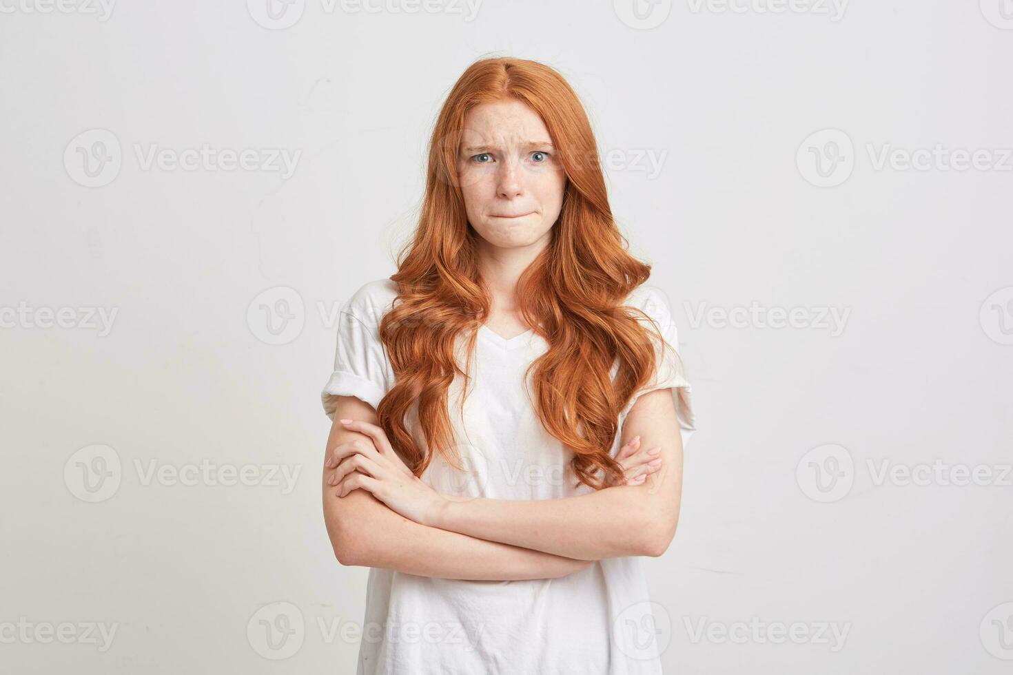 Closeup of upset worried redhead young woman with wavy long hair and freckles wears t shirt keeps hands folded and pressed her lips isolated over white background photo