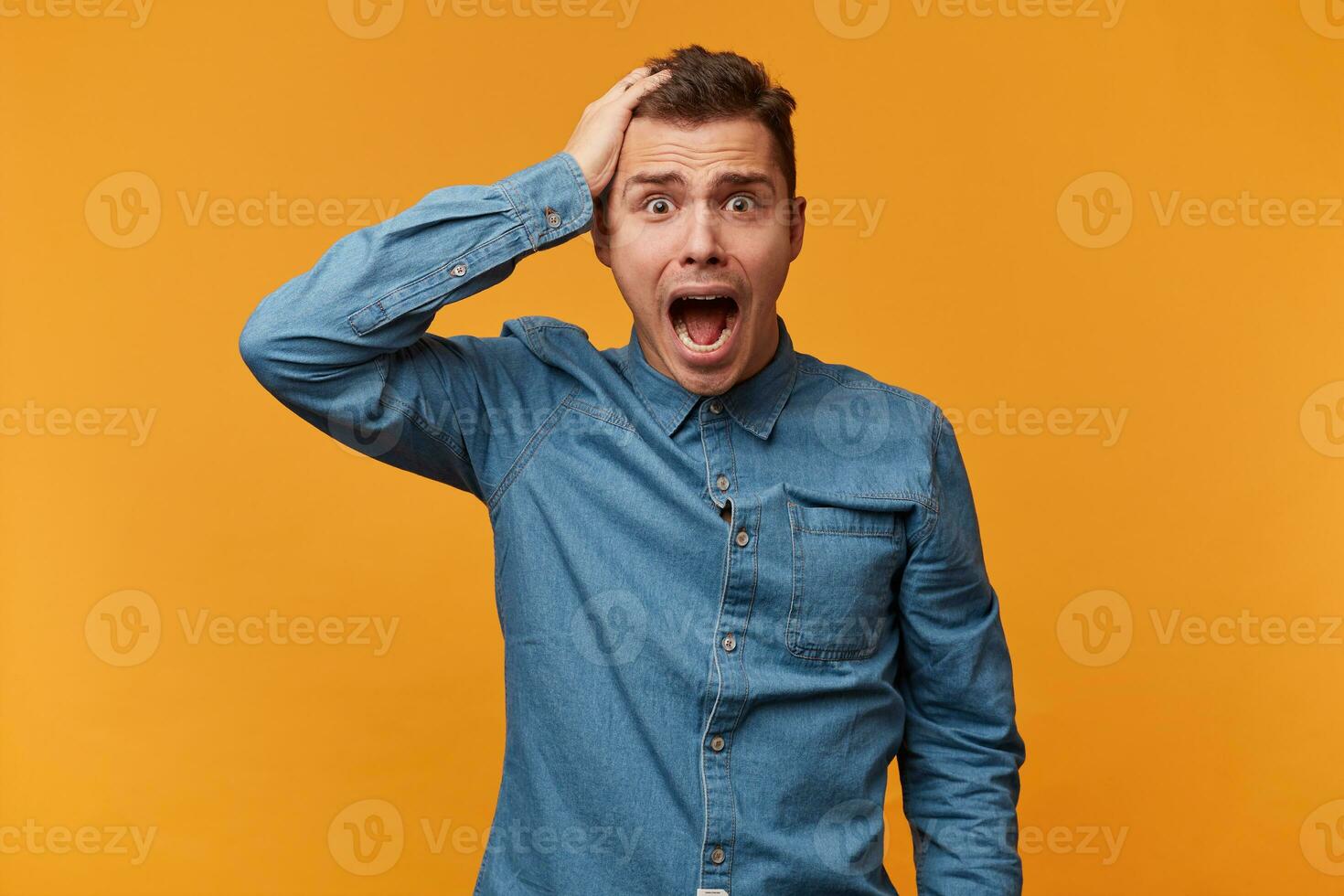 Guy in the denim shirt is holding his head with one hand, depicts a loud cry,mouth is wide open,looks confused, feels in a hopeless situation,panicking,nervous experiencing fear,over yellow background photo