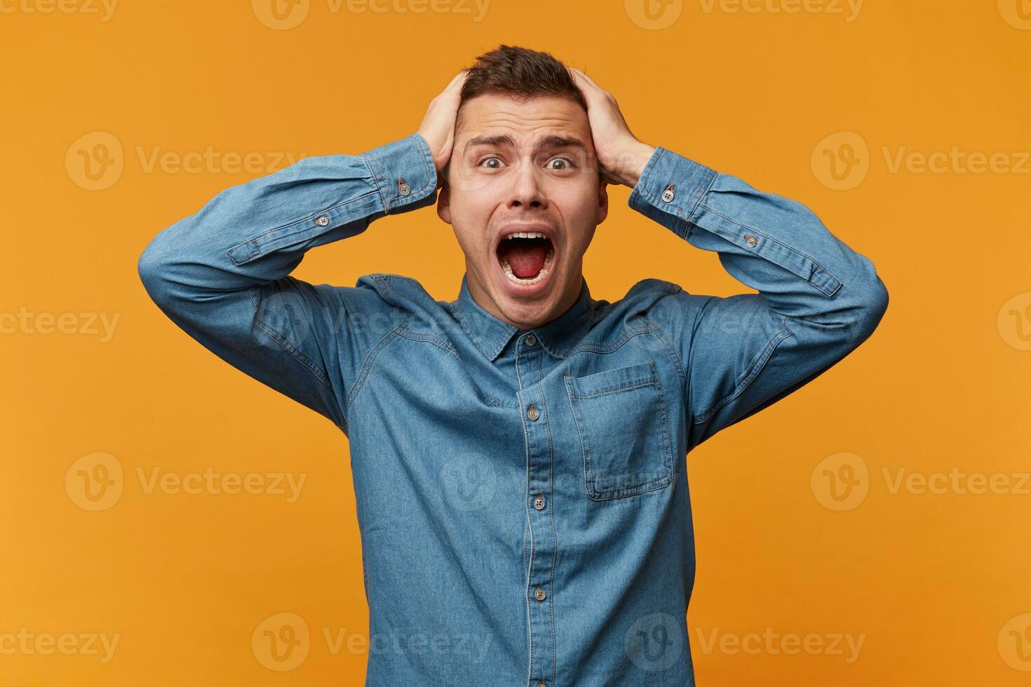 A man in a panic grabbed his head, loudly screaming, a defeat collapse fail had happened, wearing a denim shirt isolated on a yellow background. photo