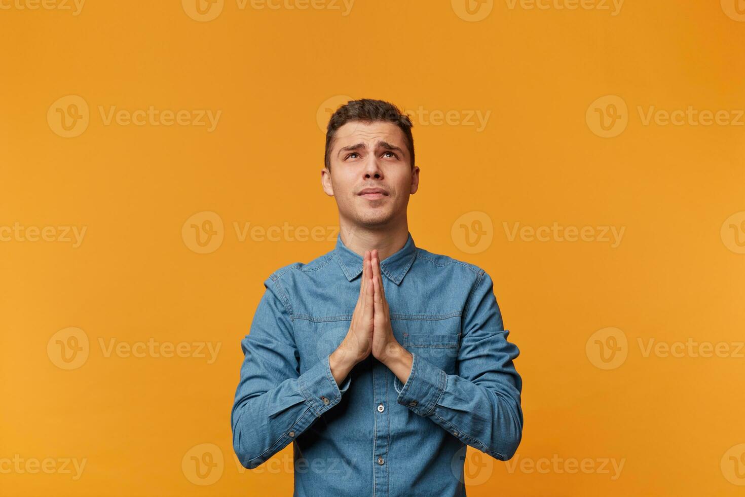 Young guy looks up with hope, palms folded in a prayer gesture, asking for help from higher powers, protection, blessing, wearing a denim shirt, isolated on a yellow background photo