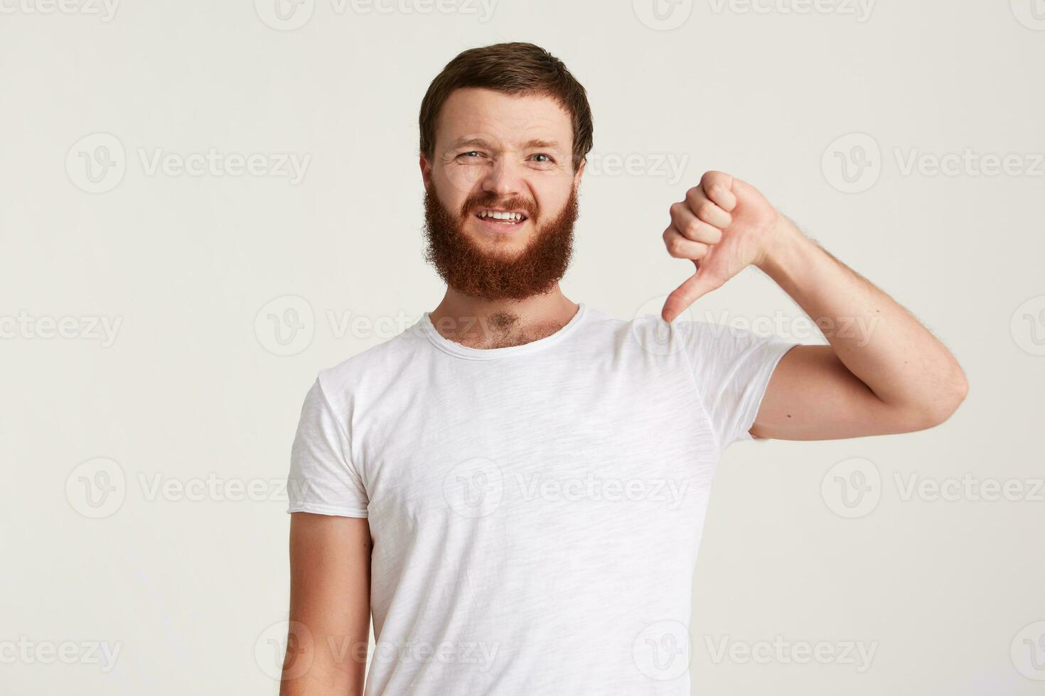 Portrait of unhappy displeased young man hipster with beard wears t shirt feels disappointed and shows thumbs down gesture isolated over white background photo