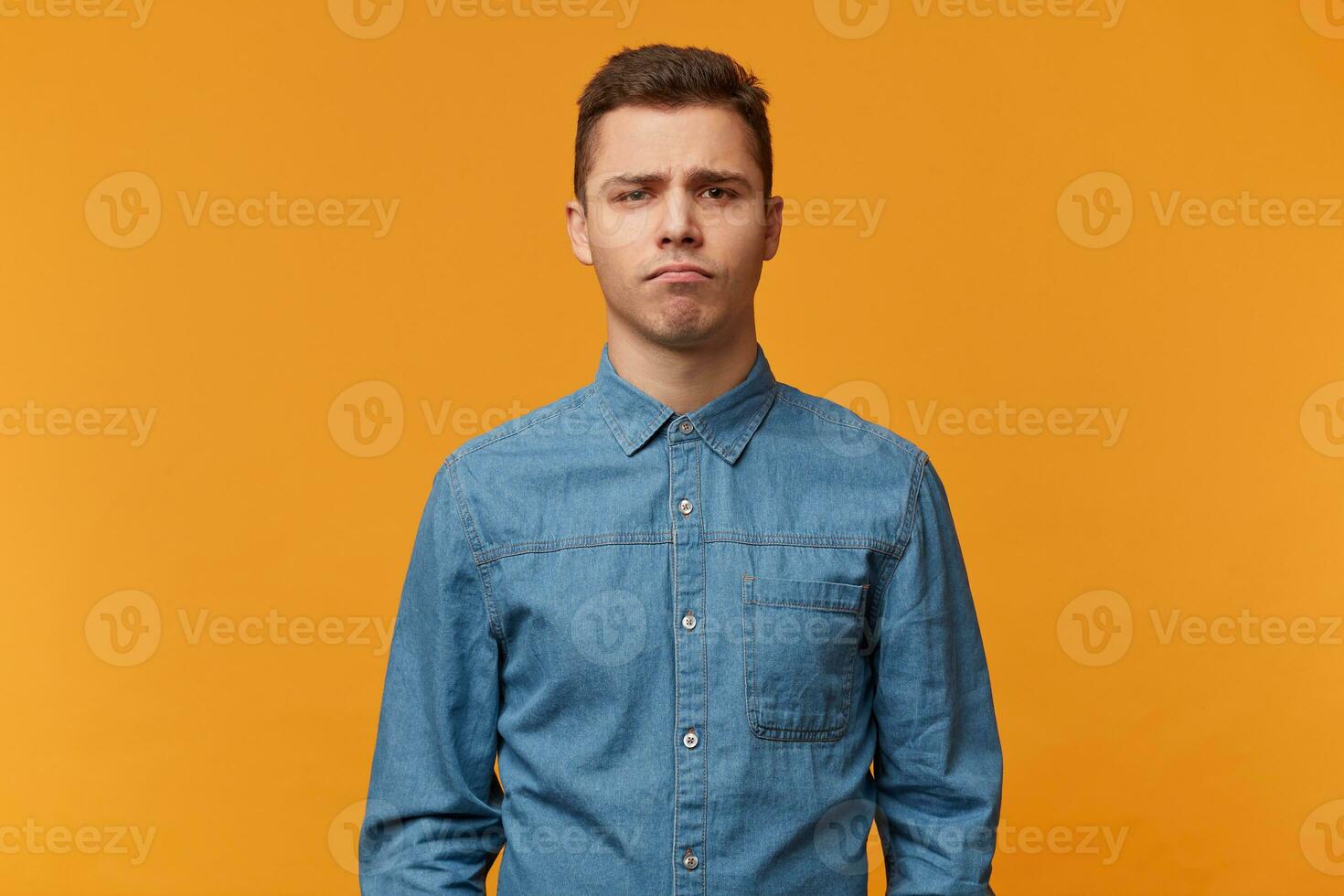 A young sad guy stands with a gloomy facial expression bored upset, isolated against a yellow background, dressed in a new denim shirt. photo