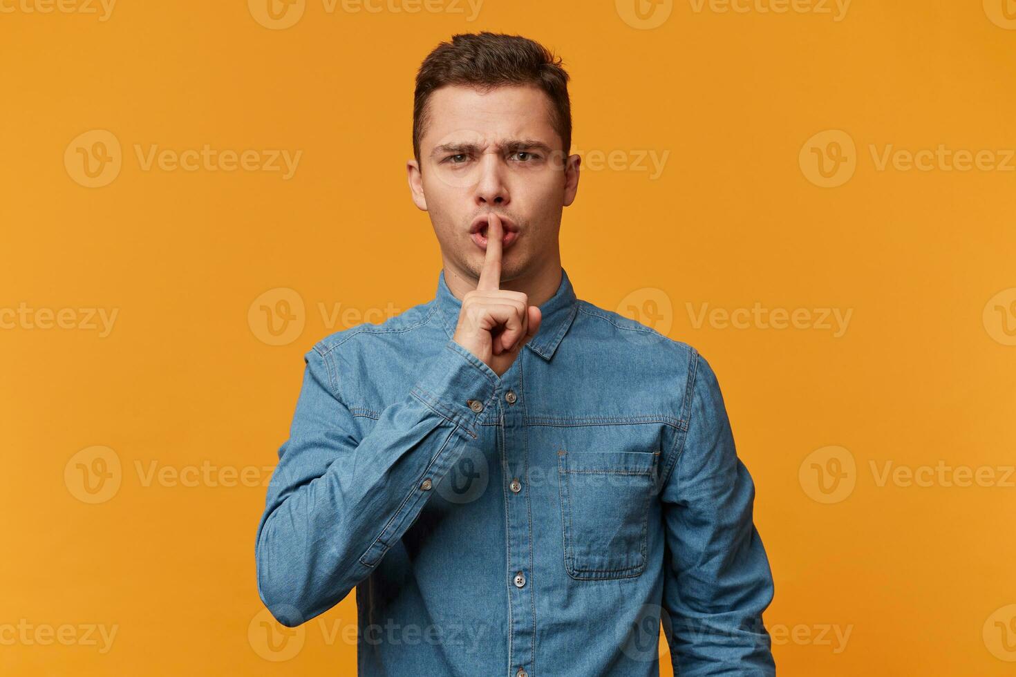Seriously looking guy in the denim shirt urges everyone to keep silence, stay calm, presses his index finger to his lips depicts a gesture of silence, over yellow background photo