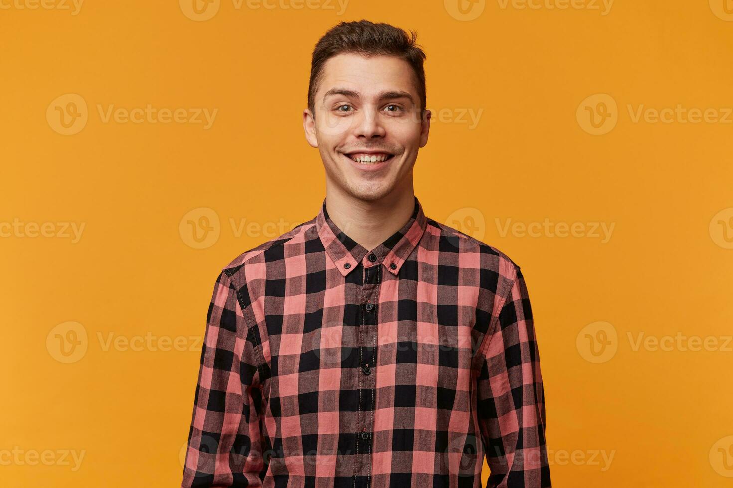 simpático atractivo alegre joven hombre sonrisas atentamente, satisfecho en un bueno ánimo, vestido en un tartán camisa, aislado en contra un amarillo antecedentes. foto