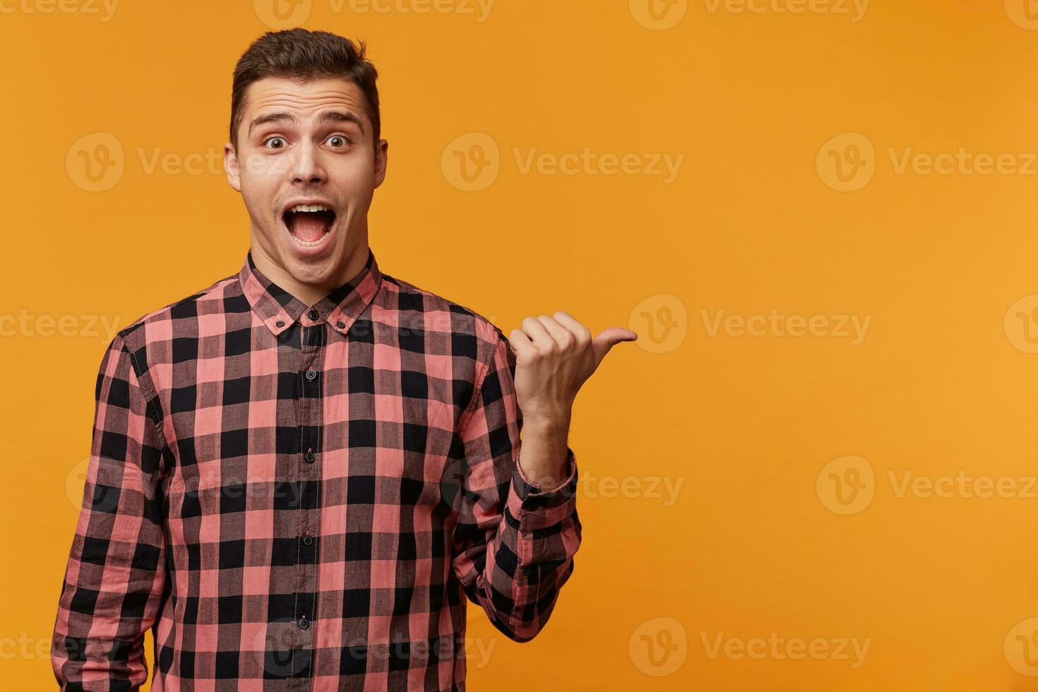 Close up of surprised amazed attractive man in denim shirt pointing rightside with dropped jaw advertise something great fantastic, standing over yellow background photo