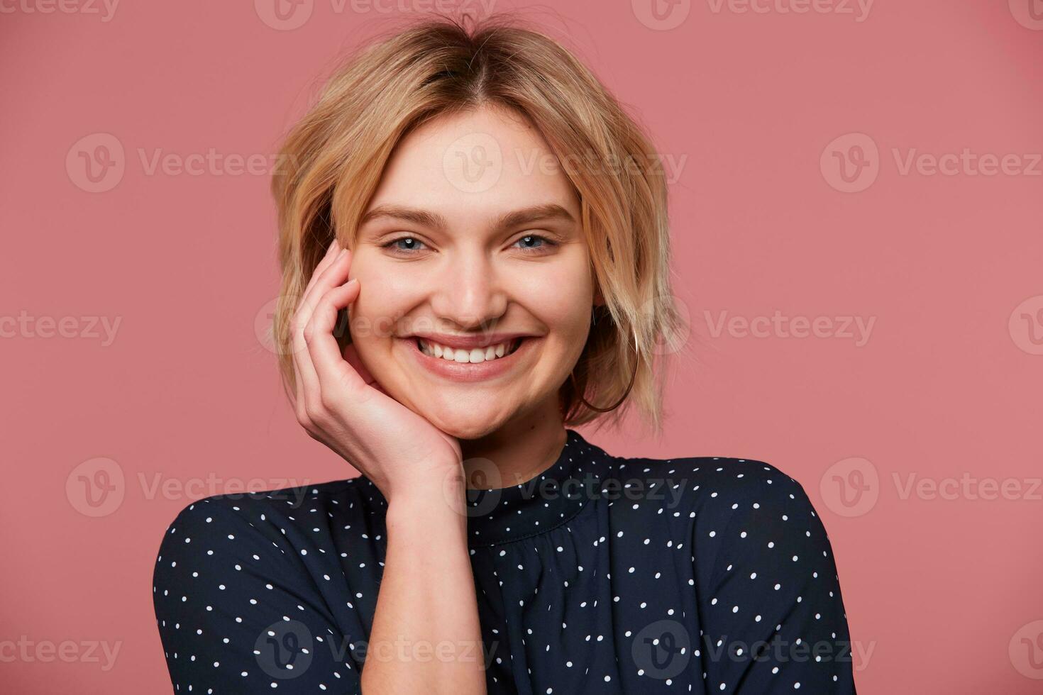 Close up of attractive joyful young beautiful blonde with short haircut dressed in blouse with polka dots, coquets, flirting, smiling pleasantly, isolated over pink background photo