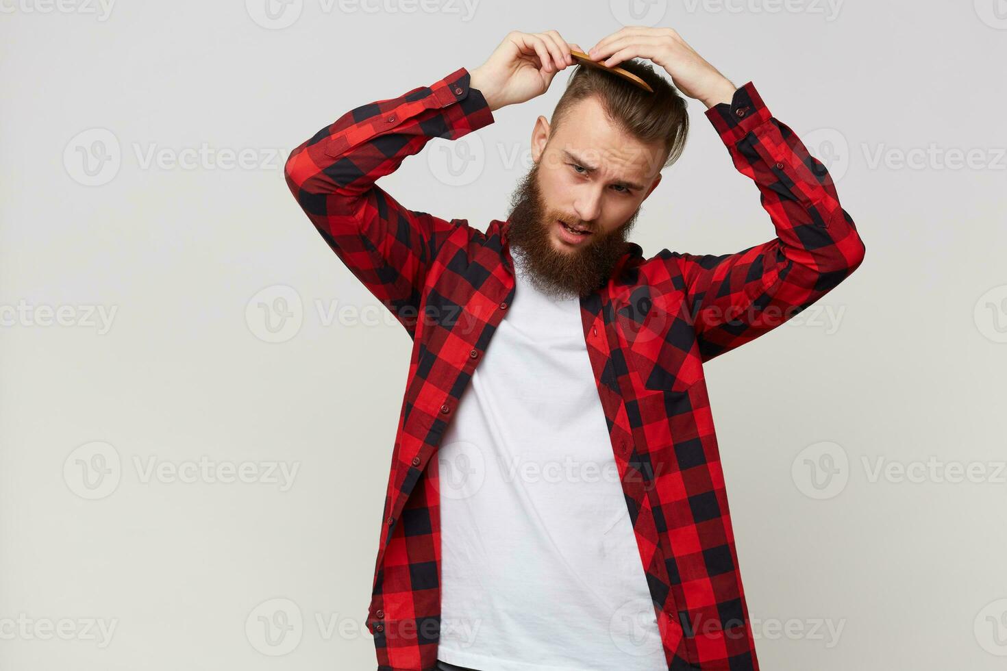 Bearded man in shirt with not very glad facial expression doing modern hairstyle, grooming his hair with comb with discontent isolated over white background photo