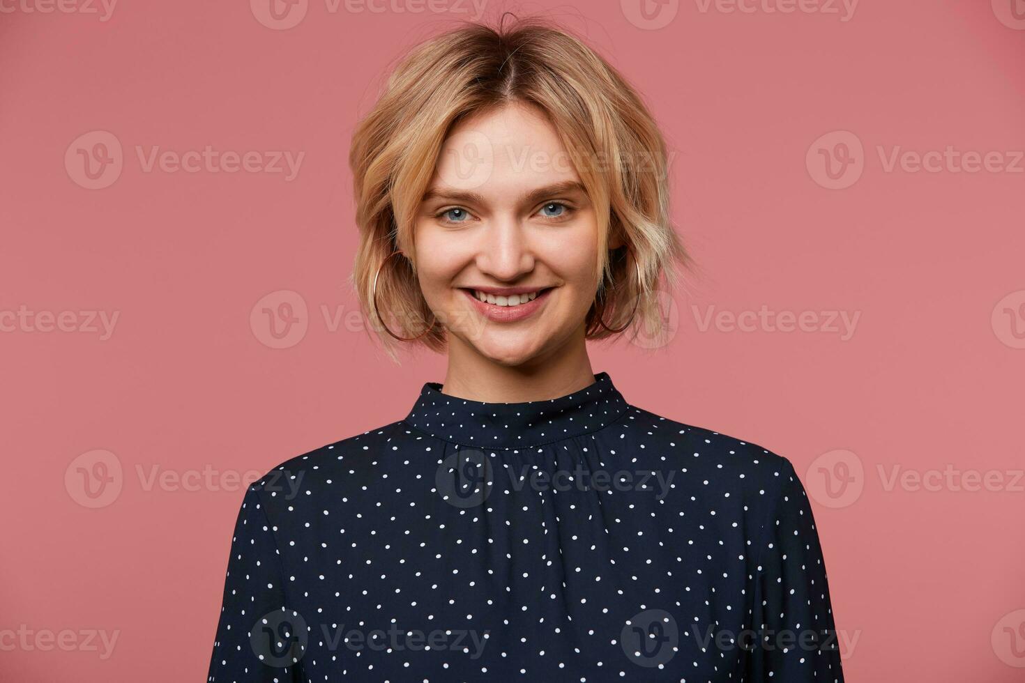 Pleasantly smiling young beautiful attractive blonde woman isolated over pink background dressed in blouse with polka dots, has glad face expression, showing positive, happiness photo