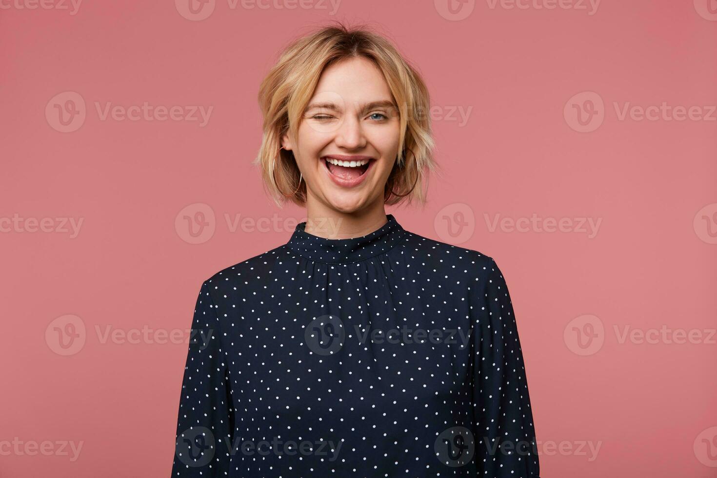 Playful young beautiful attractive blonde woman dressed in blouse with polka dots, winks, showing positive, flirting, smiling, happy, isolated over pink background photo