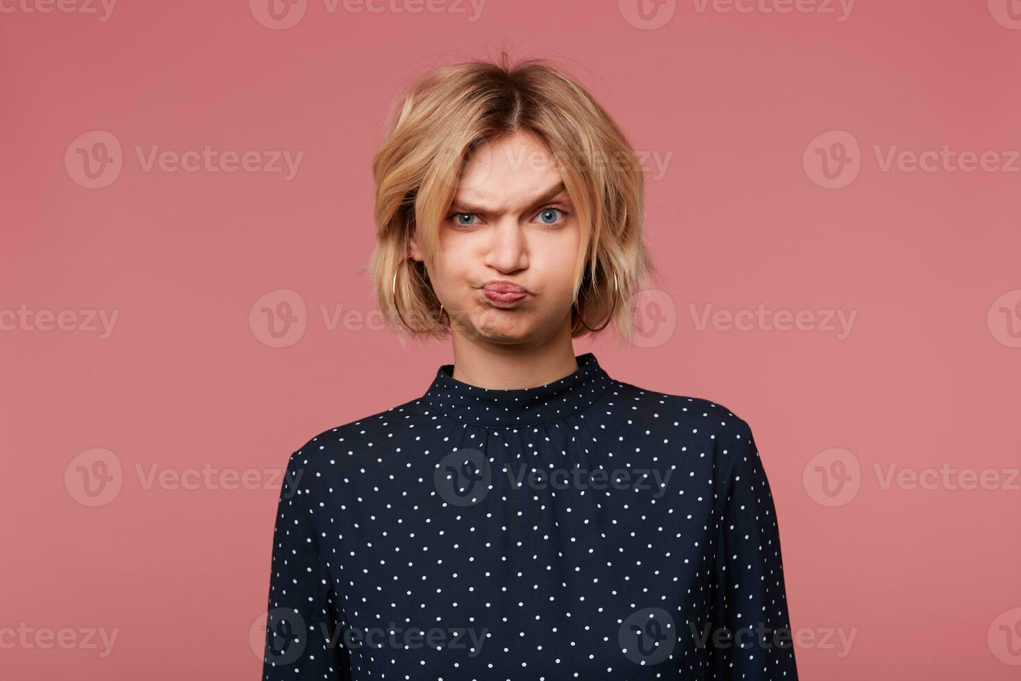 Blonde girl offended angry upset, pouted her cheeks is overwhelmed with negative emotions, in bad mood dressed in blouse with polka dots,isolated on pink photo