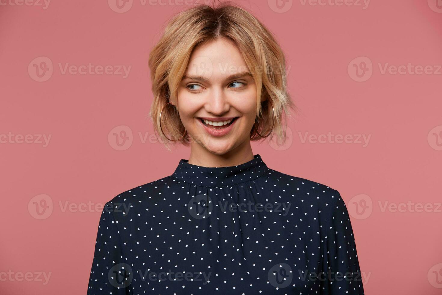 Nice young beautiful attractive blonde woman dressed in blouse with polka dots, has glad face expression, smiling, talking coquettishly looking to the side, isolated over pink background photo