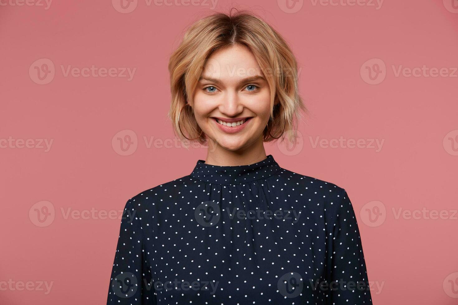 estudio Disparo de sonriente hermosa atractivo rubia mujer con corto Corte de pelo aislado terminado rosado antecedentes vestido en blusa con polca puntos, tiene alegre cara expresión, demostración positivo, felicidad foto