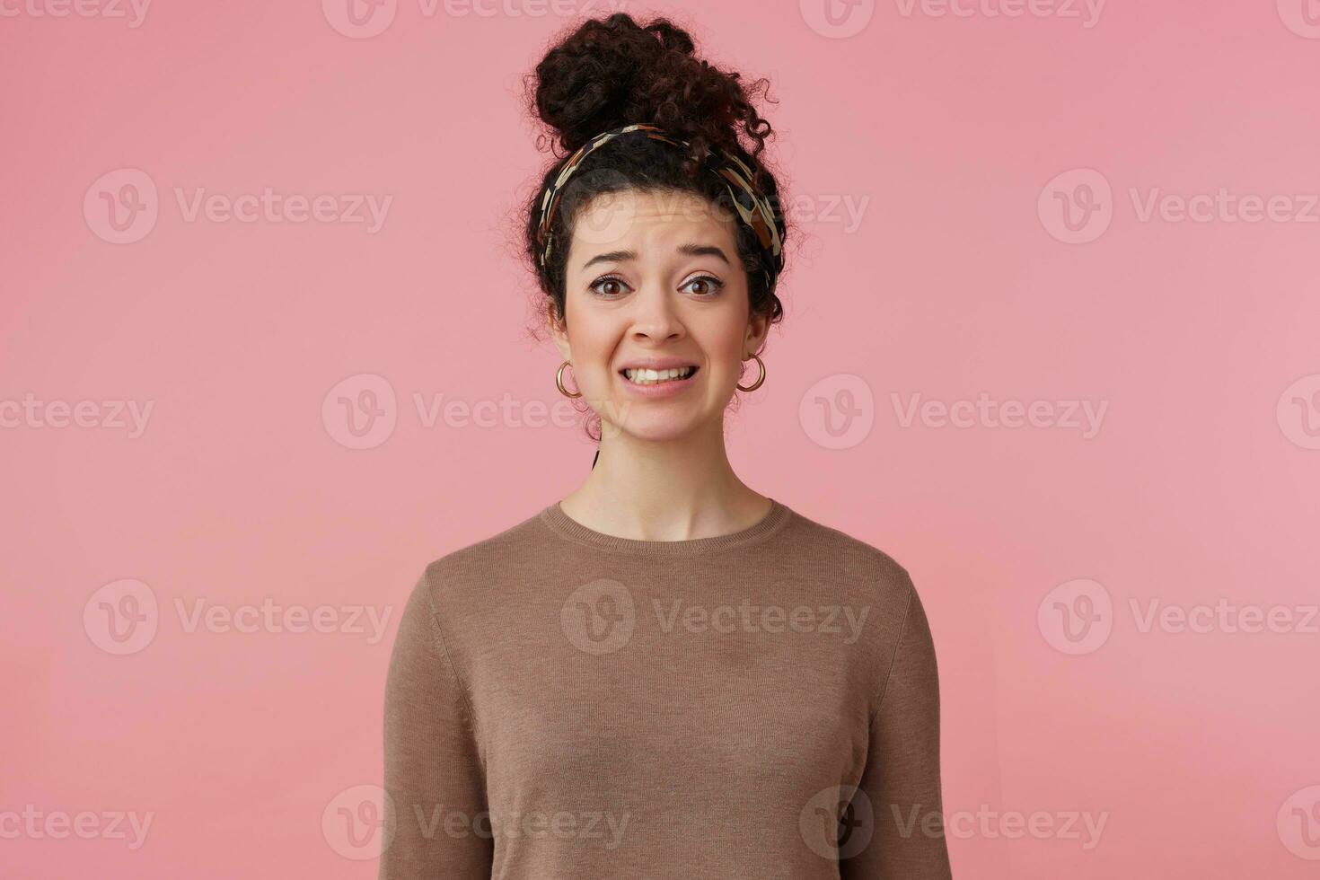 Discontent woman, unsure girl with dark curly hair bun. Wearing headband, earrings and brown sweater. Has make up. Emotion concept. Watching at the camera isolated over pastel pink background photo