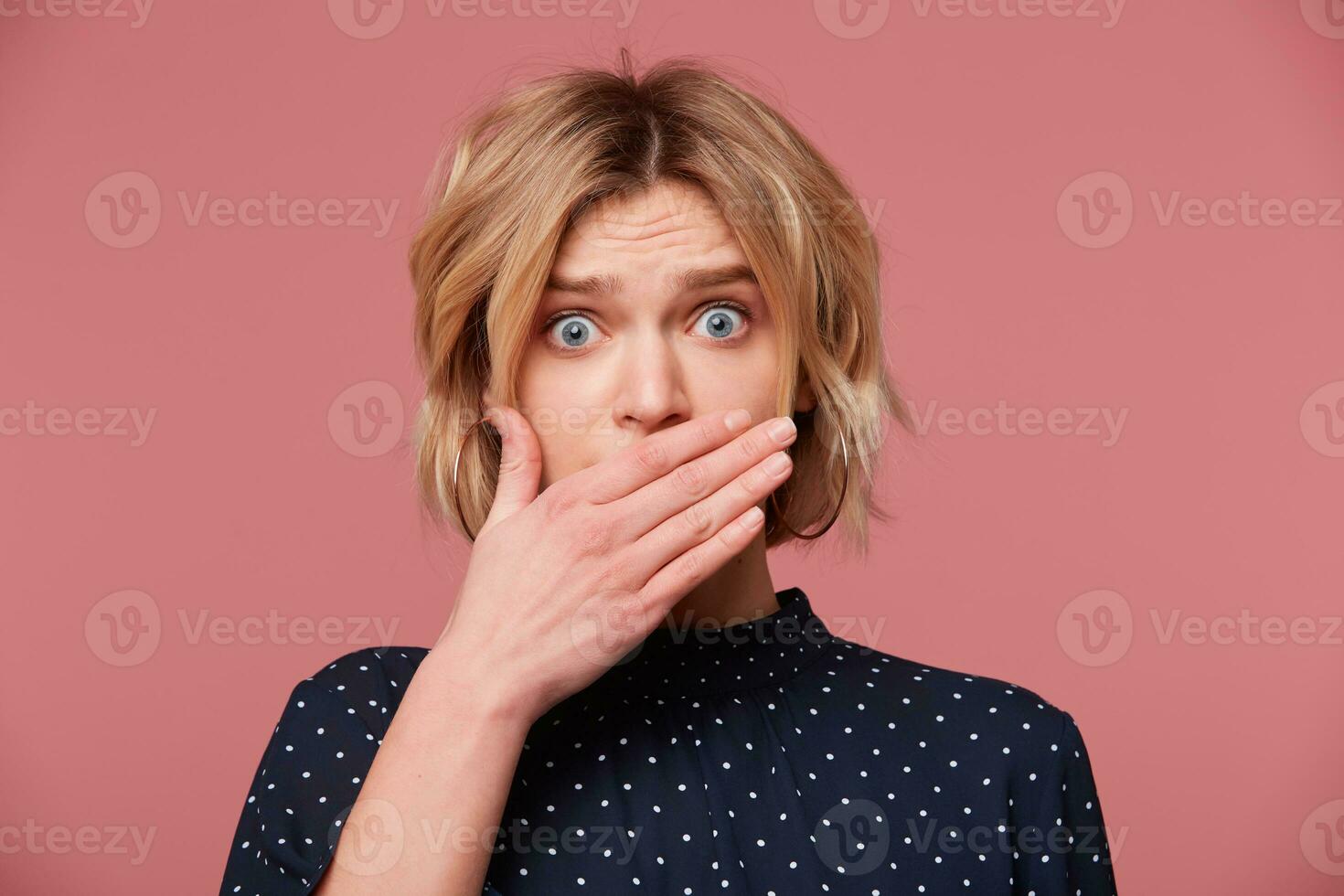 Portrait of irresponsible woman having troubles. Frightened blonde in despair covered mouth with palm, scares to talk, dressed blouse with polka dots, isolated over pink background photo