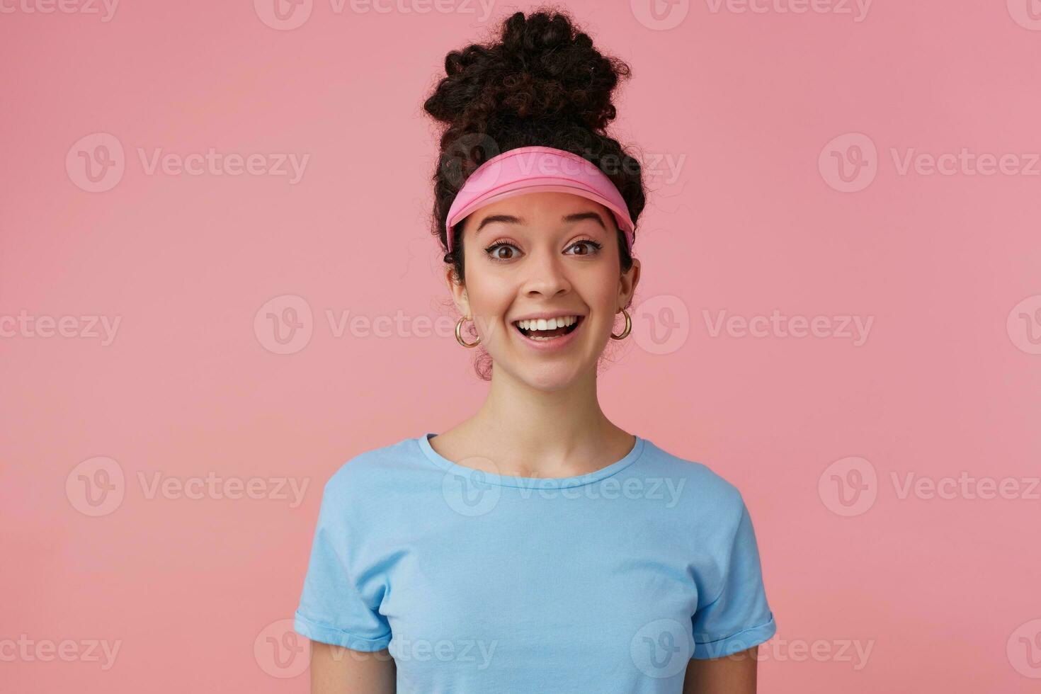 positivo chica, contento mirando mujer con oscuro Rizado pelo bollo. vistiendo rosado visera, pendientes y azul camiseta. tiene hacer arriba. emoción concepto. acecho a el cámara aislado terminado pastel rosado antecedentes foto