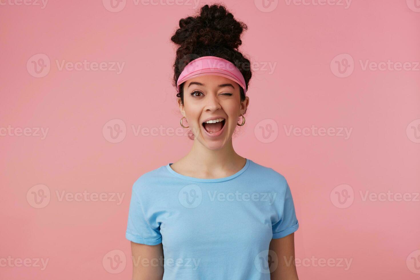 Portrait of attractive, playful girl with dark curly hair bun. Wearing pink visor, earrings and blue t-shirt. Has make up. Emotion concept. Winks at the camera isolated over pastel pink background photo