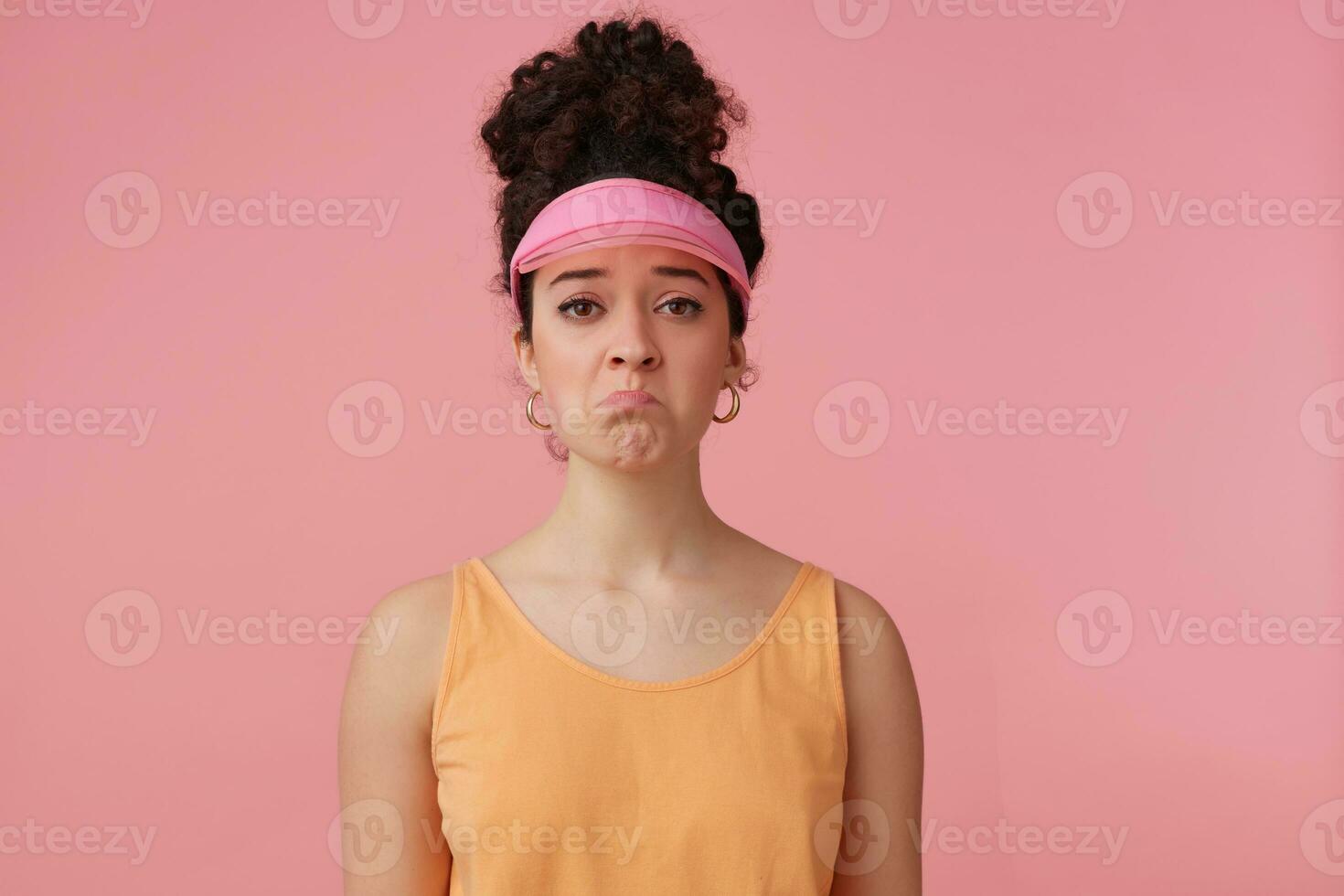 Portrait of sad girl with dark curly hair bun. Wearing pink visor, earrings and orange tank top. Has make up. Emotion concept. Pouts a lip. Watching at the camera isolated over pastel pink background photo