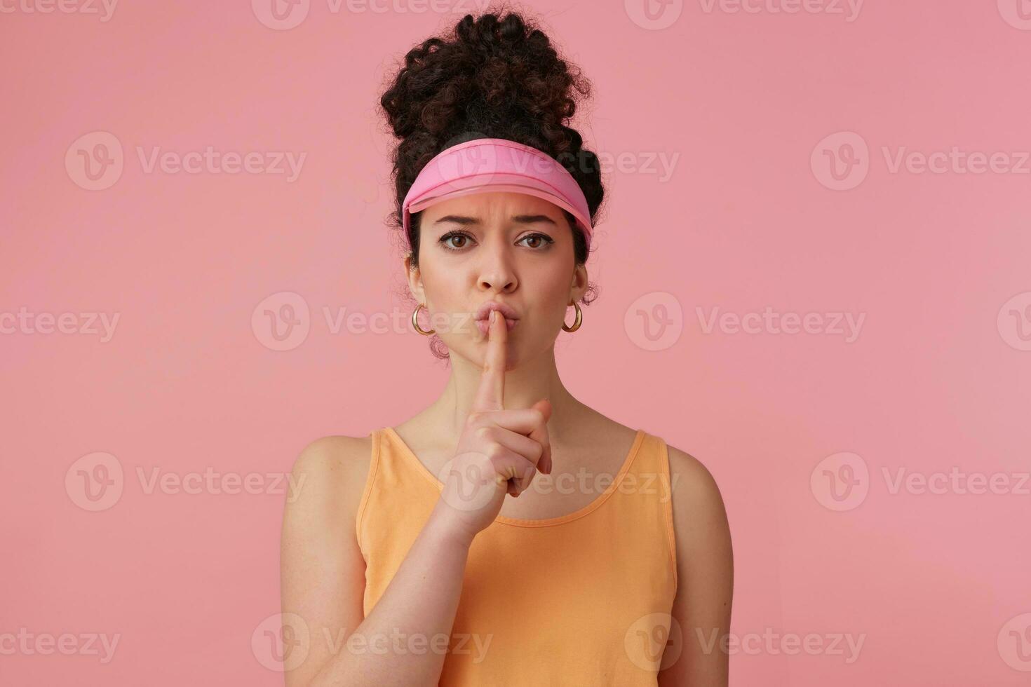 Frowning woman, beautiful girl with dark curly hair bun. Wearing pink visor, earrings and orange tank top. Has make up. Shows silence sign. Watching at the camera isolated over pastel pink background photo