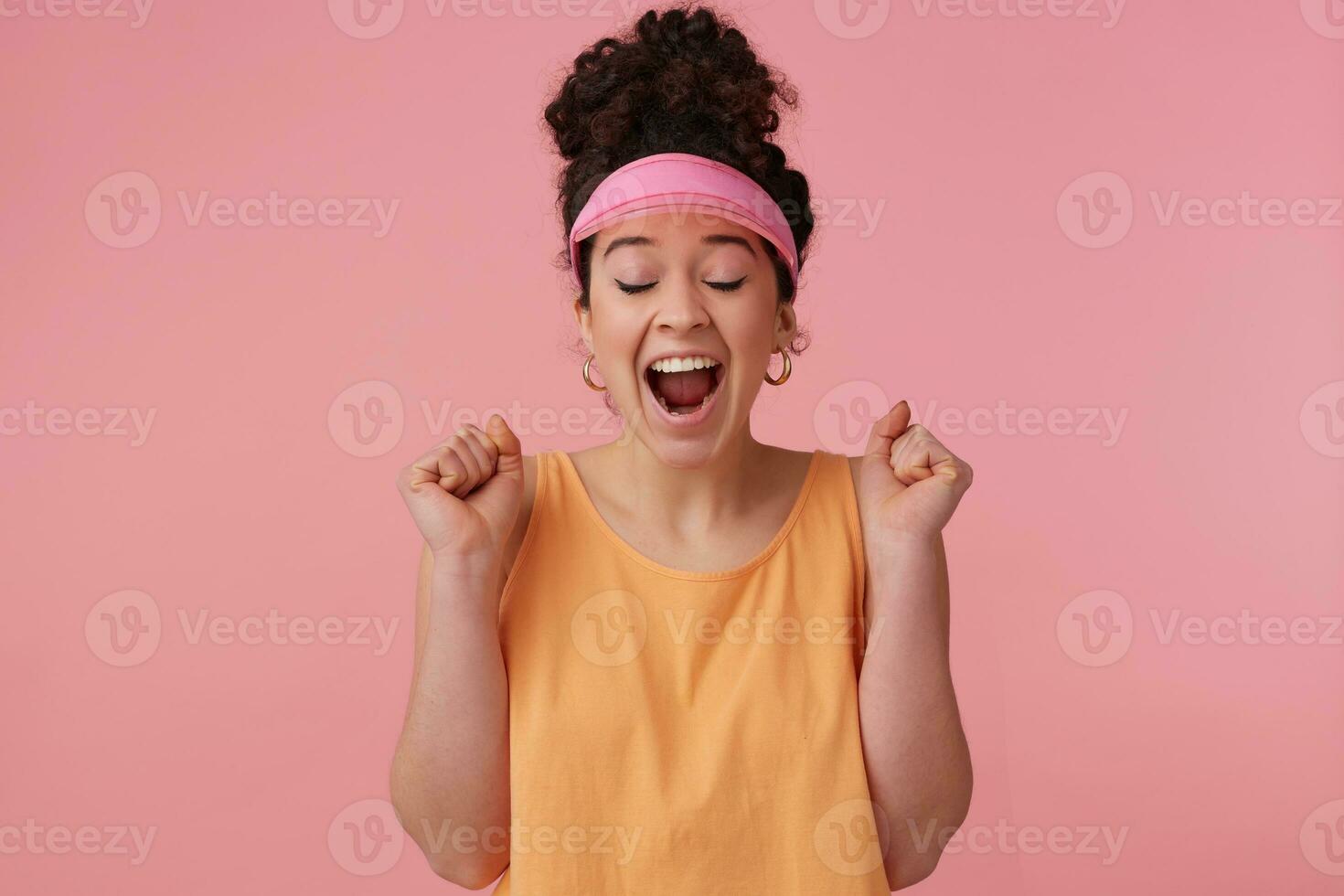 alegre, sorprendido niña con oscuro Rizado pelo bollo. vistiendo rosado visera, pendientes y naranja tanque cima. tiene hacer arriba. apretar puños y cerca ojos en excitación. estar aislado terminado pastel rosado antecedentes foto