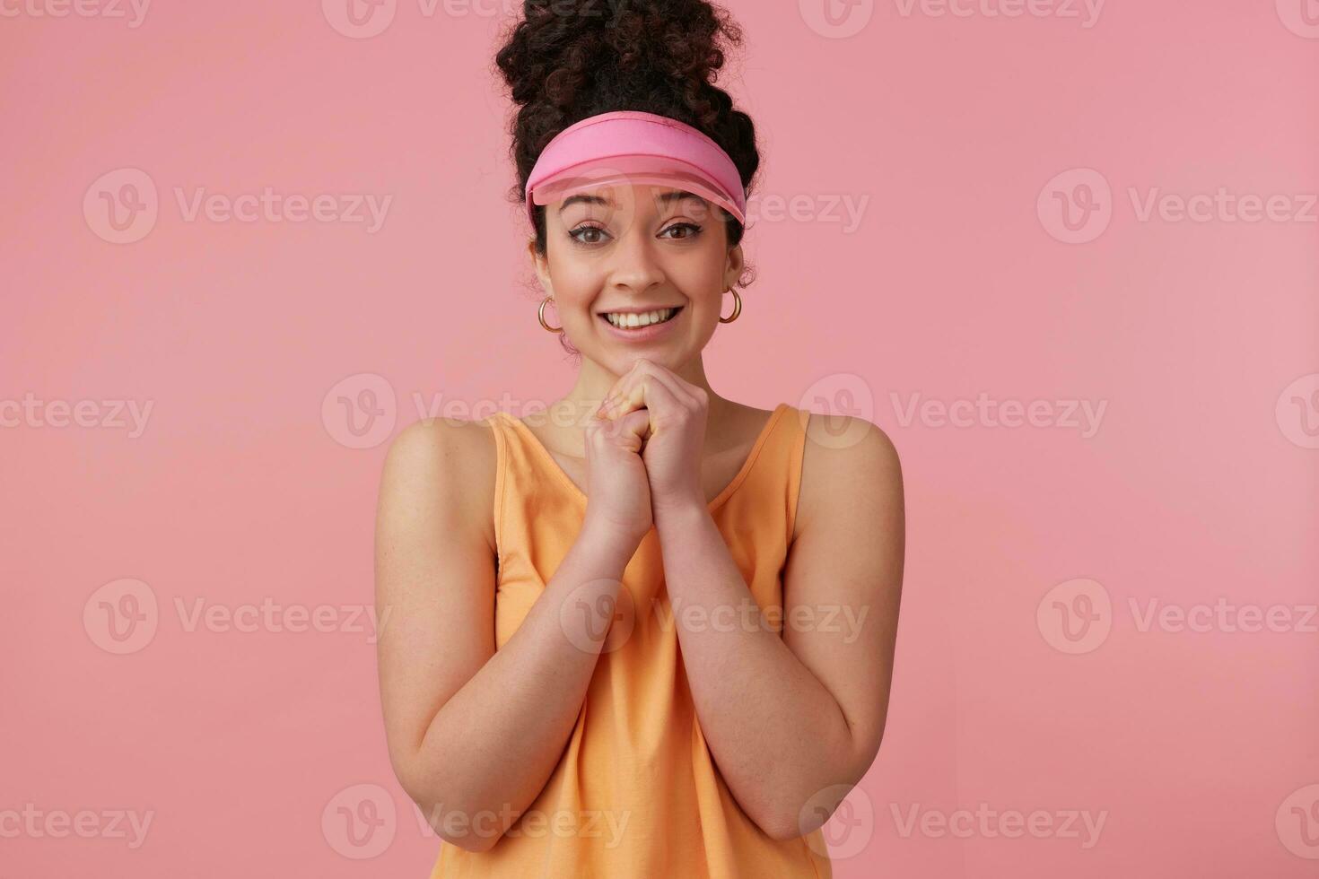 Portrait of pleading girl with dark curly hair bun. Wearing pink visor, earrings and orange tank top. Has make up. Keeps palms together. Watching at the camera isolated over pastel pink background photo