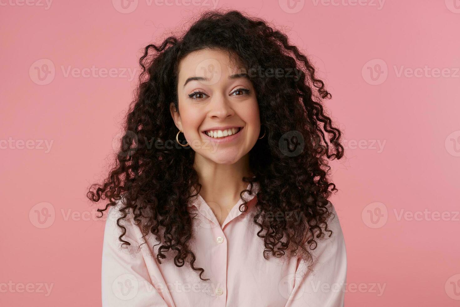 Portrait of attractive, adult girl with long dark curly hair. Wearing earrings and pastel pink shirt. Has make up. Broadly smiling. Watching at the camera isolated over pastel pink background photo