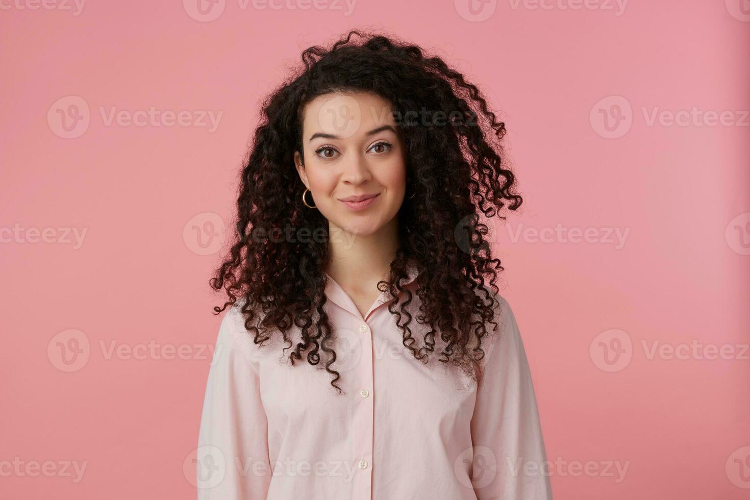 Charming girl, happy looking woman with dark long hair. Wearing earrings and pastel pink shirt. Has make up. People and emotion concept. Watching at the camera isolated over pastel pink background photo