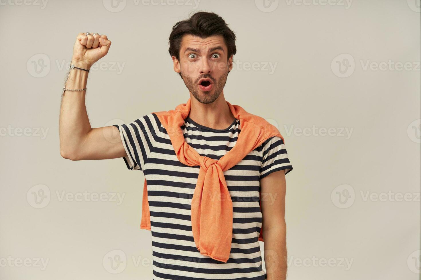 Rebellious guy with dark hair and bristle. Wearing striped t-shirt, sweater tied on shoulders. Raises fist up, protesting. Has bracelets and rings. Watching at the camera isolated over grey background photo