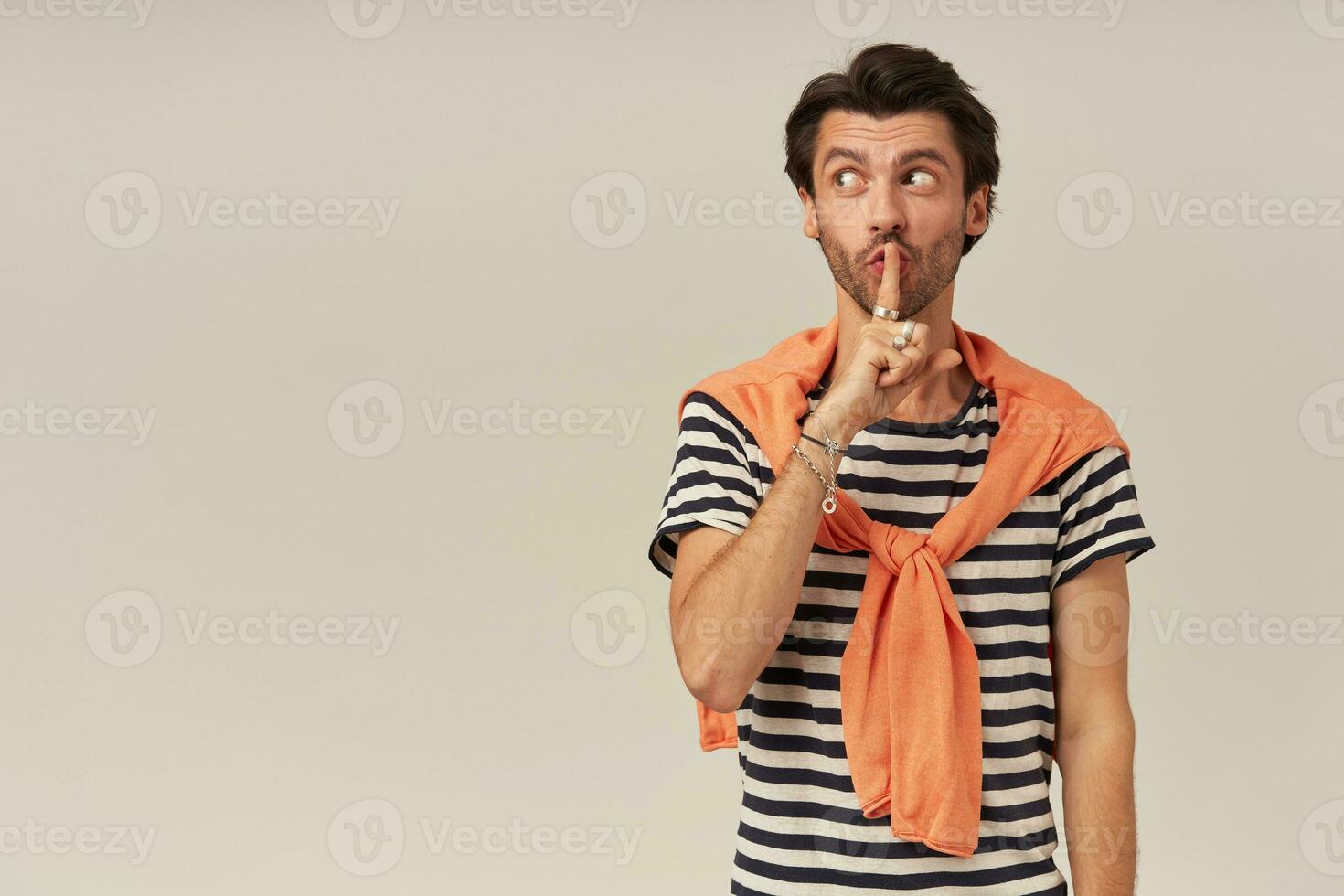 Amazed man with brunette hair and bristle. Wearing striped t-shirt and orange sweater tied on shoulders. Showing silence sign. Watching to the left at copy space, isolated over grey background photo