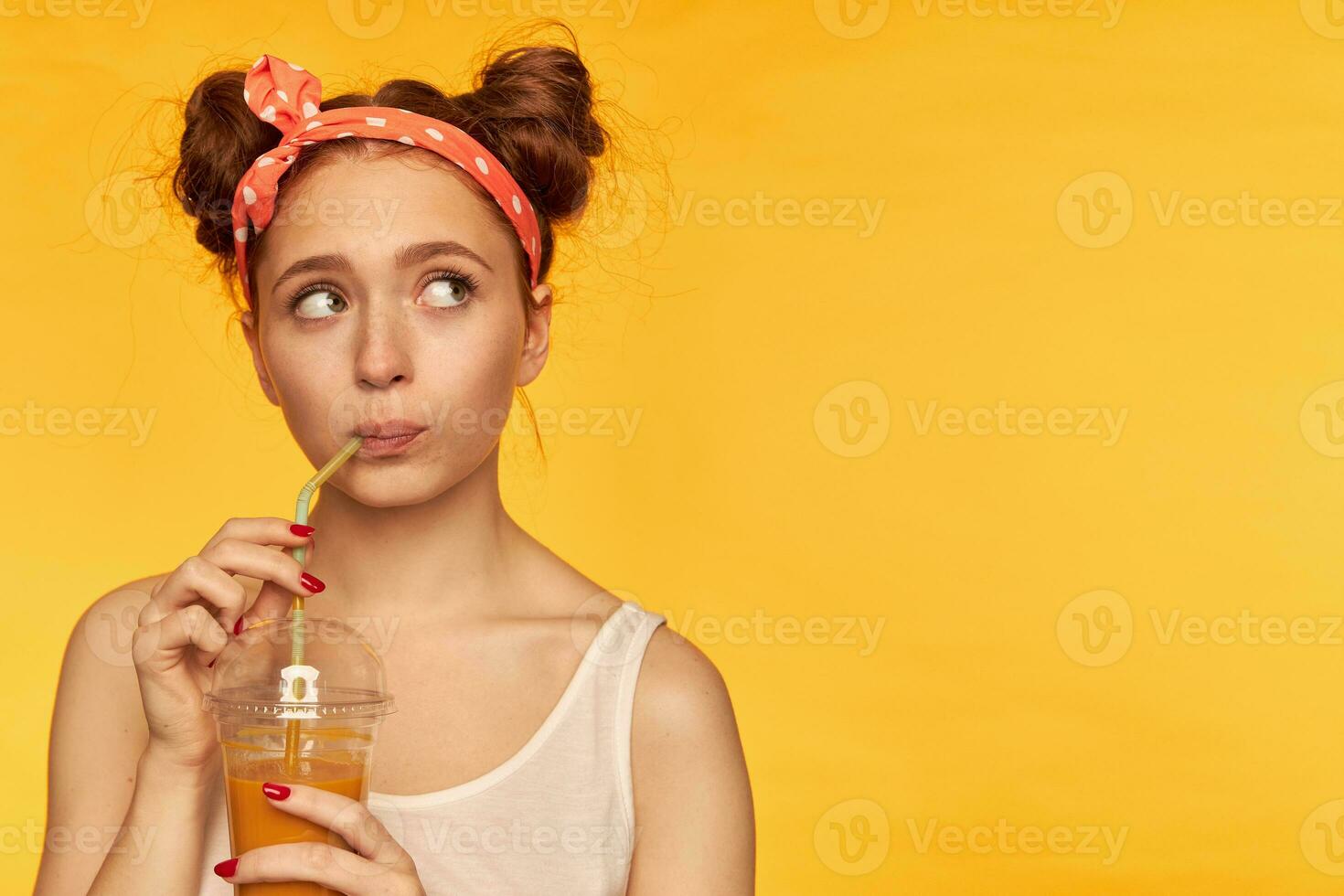 Young lady, pretty ginger woman with two buns and doted hairband. Wearing white shirt and wonder while making a sip of juicy fresh. Watching to the right at copy space over yellow background photo