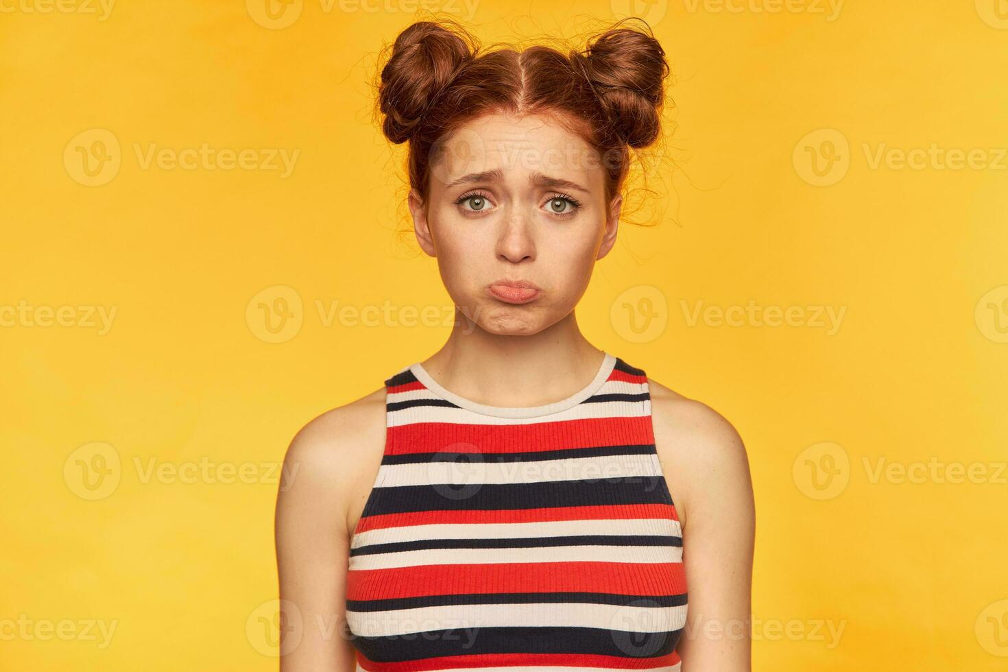 Young lady, unhappy ginger woman with two buns. Wearing striped tank top and looking sad, you offend her. Emotional concept. Watching at the camera isolated over yellow background photo