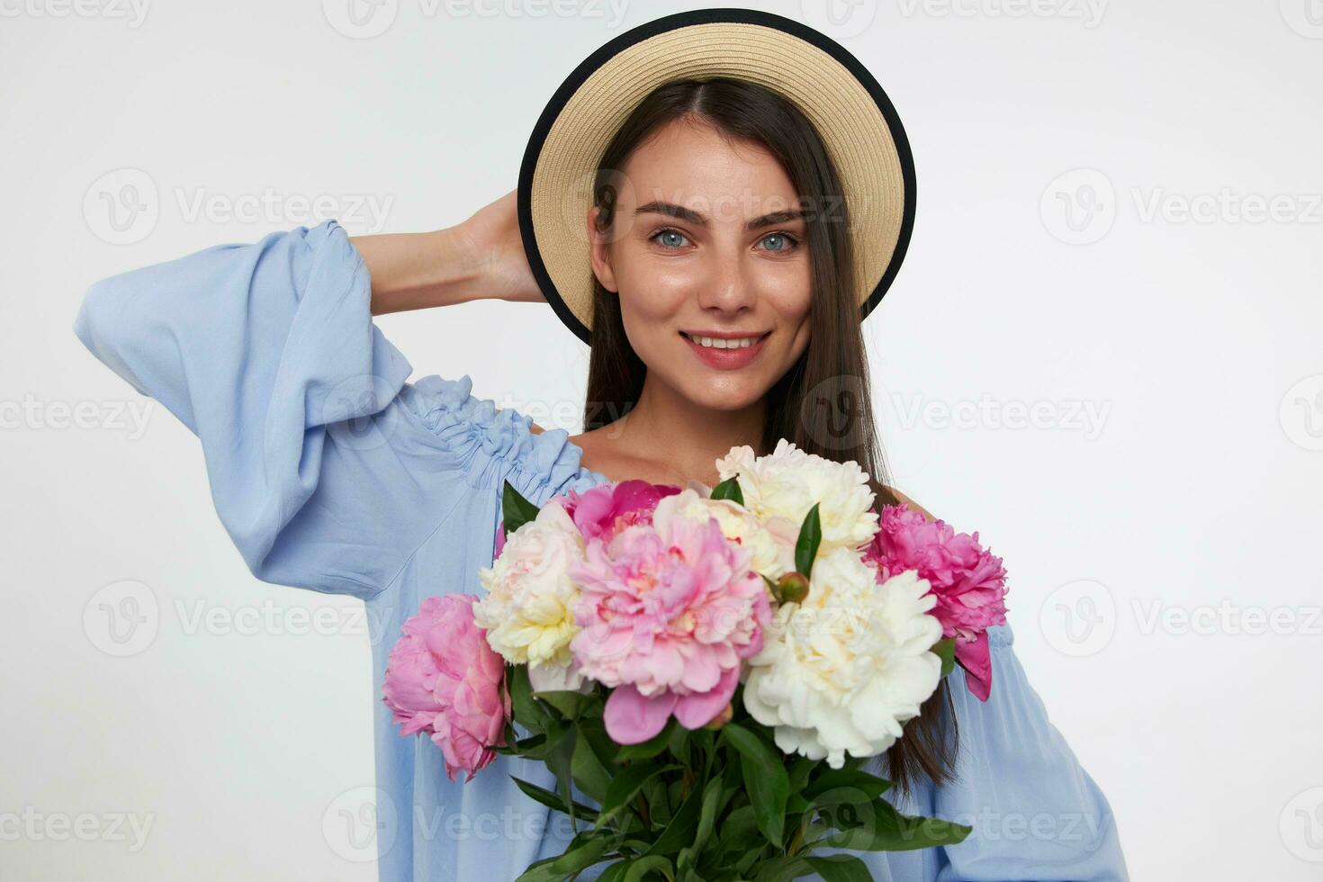 Teenage girl, happy looking woman with brunette long hair. Wearing a hat and blue dress. Holding bouquet of flowers and touching her head. Watching at the camera isolated over white background photo