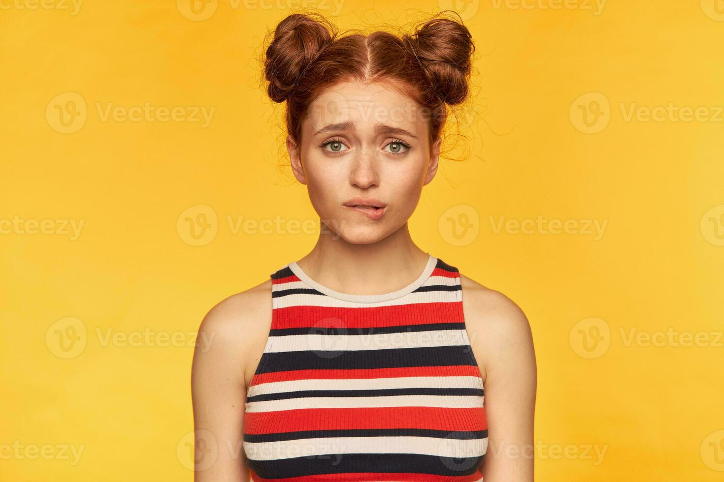 Teenage girl, happy looking red hair woman with two buns. Wearing striped tank top and bite her lip, pending of what you will say. Watching at the camera isolated over yellow background photo