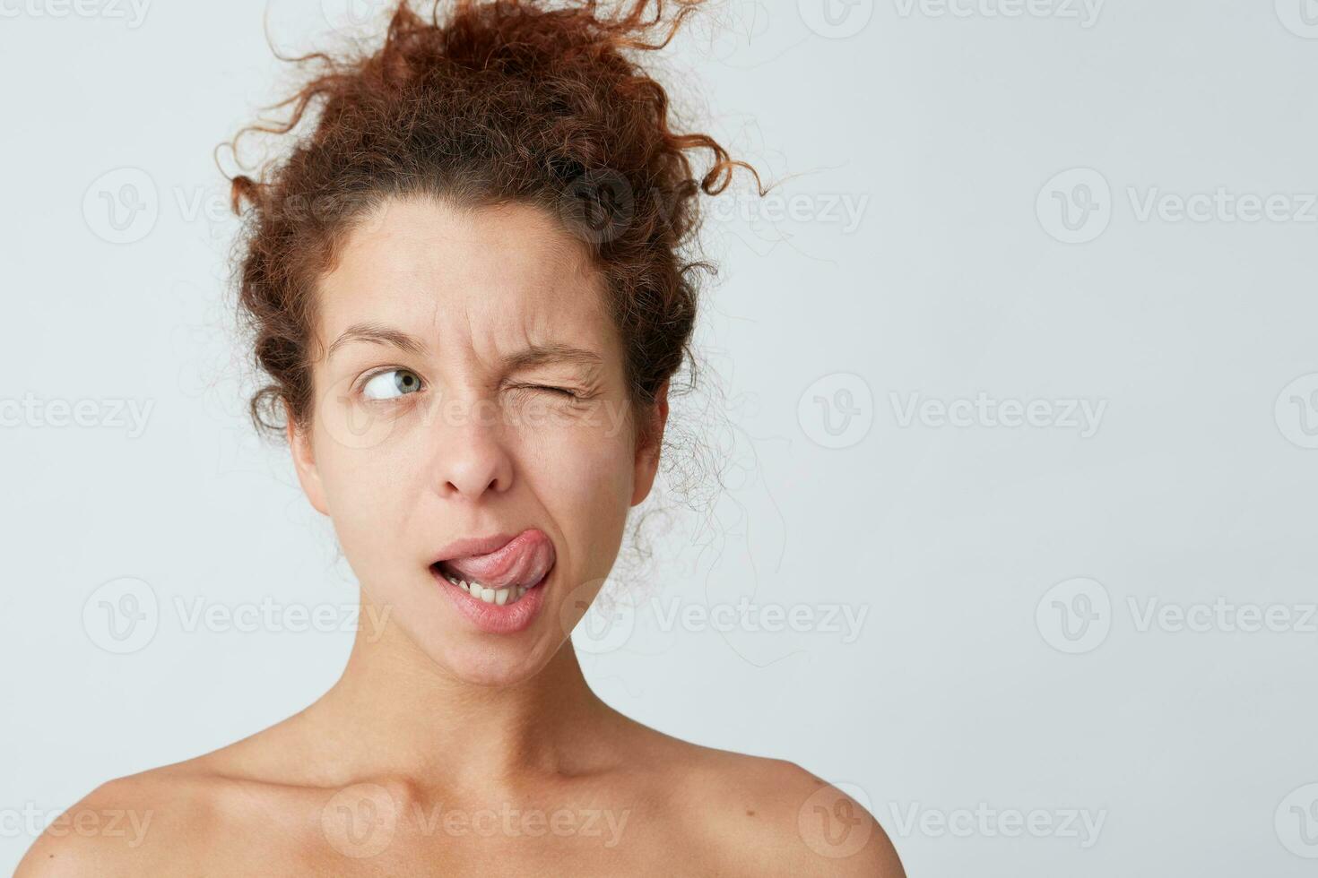 Close up shot of thoughtful playful young woman with curly hair and perfect skin shows tongue, winks and looks to the side isolated over white background Thinking about her job before taking shower photo