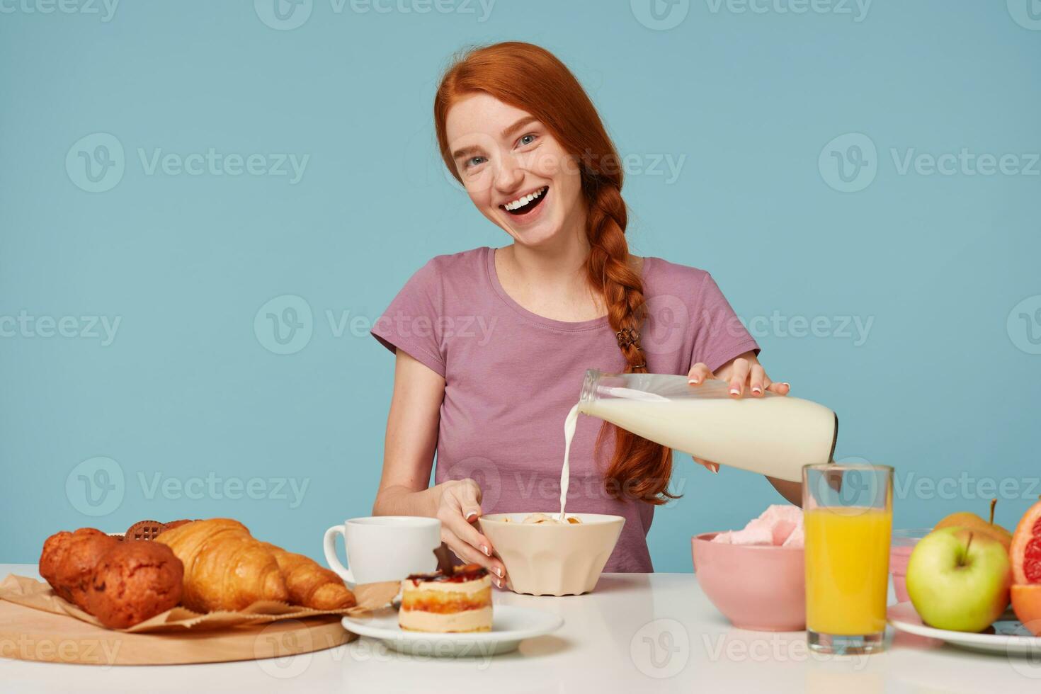 Pretty red haired girl has a good healthy breakfast,puts milk to a cereals, happyly smiling isolated on blue background photo