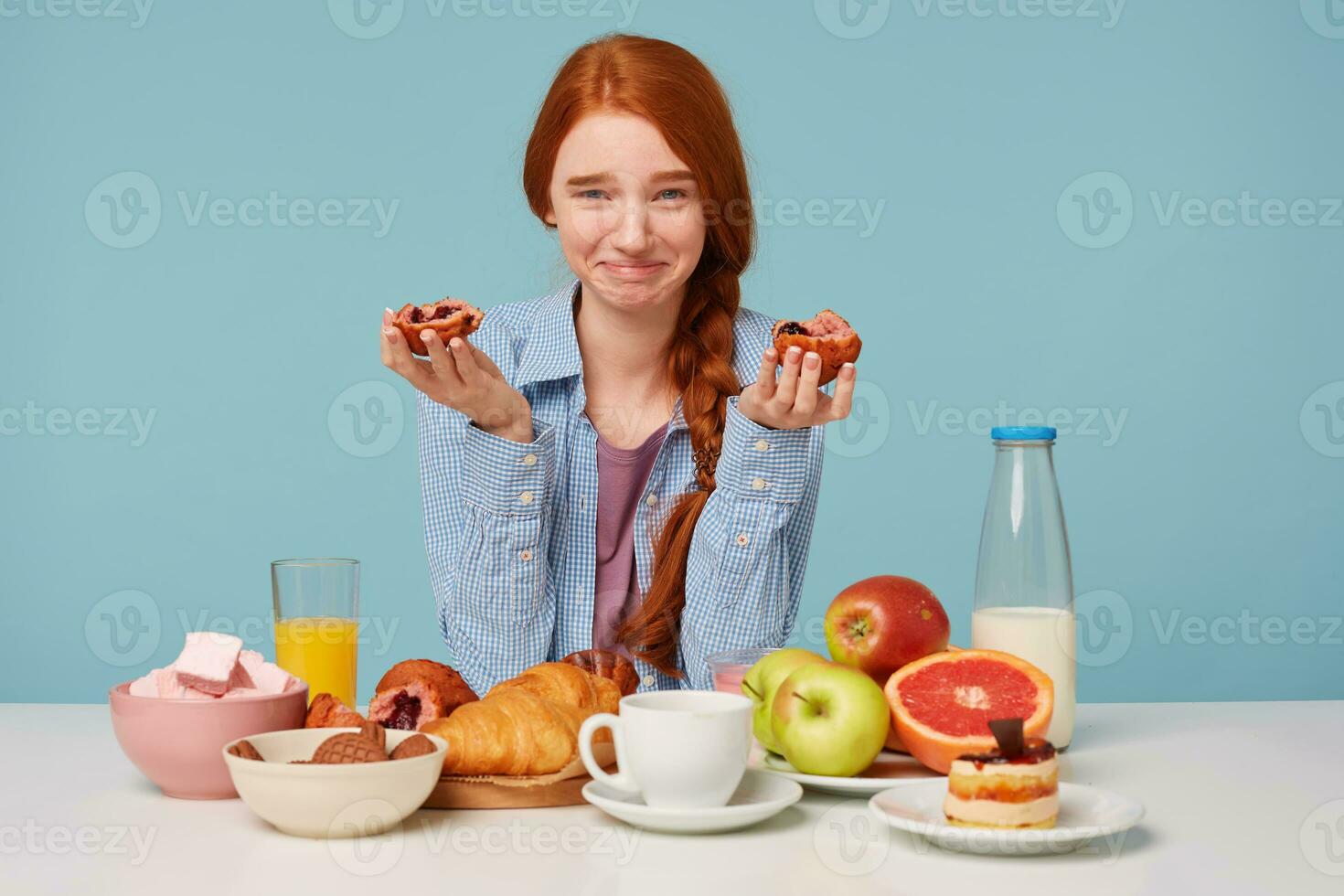 Red-haired happy funny girl has breakfast, broke off two slices of bun keeps them in hands and laughing with closed mouth,isolated on blue background photo