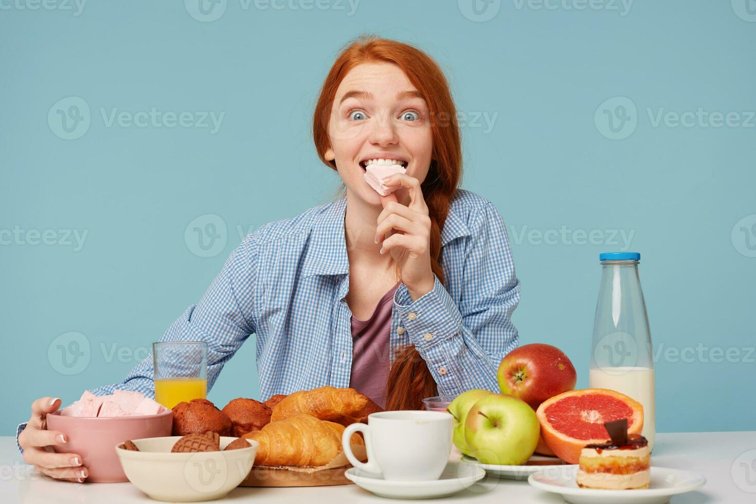 Portrait of an excited red haired girl holding marshmallow having a variable breakfast and looking at camera isolated over blue background photo