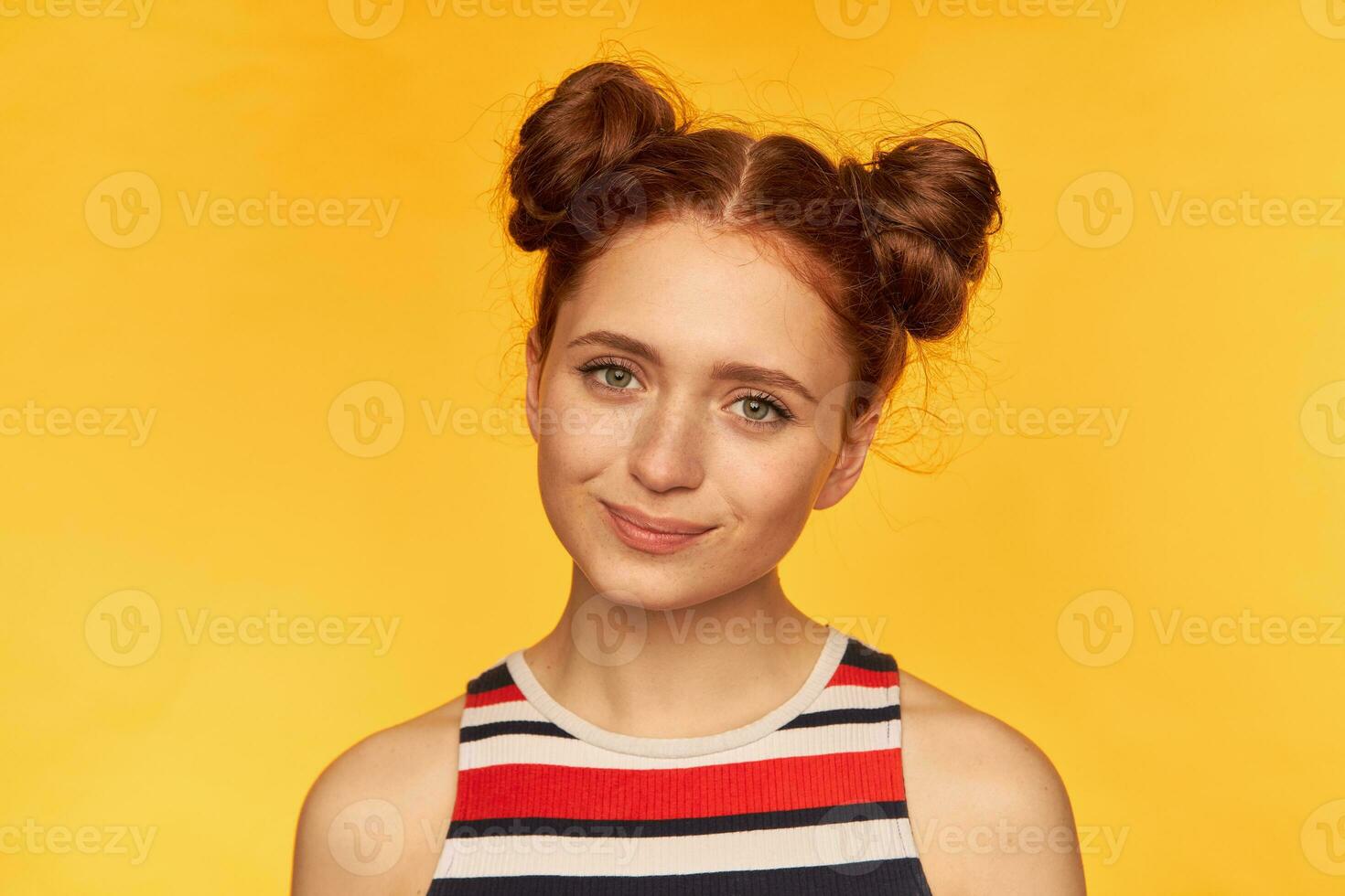 retrato de atractivo, rojo pelo niña con dos bollos y sano piel. vistiendo a rayas camisa y acecho a el cámara con linda sonrisa, de cerca, estar aislado terminado amarillo antecedentes foto