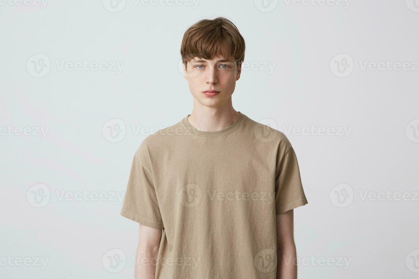 Portrait of serious handsome young man student with short haircut and blond hair in beige t shirt standing and looking directly in camera isolated over white background Feels concentrated photo