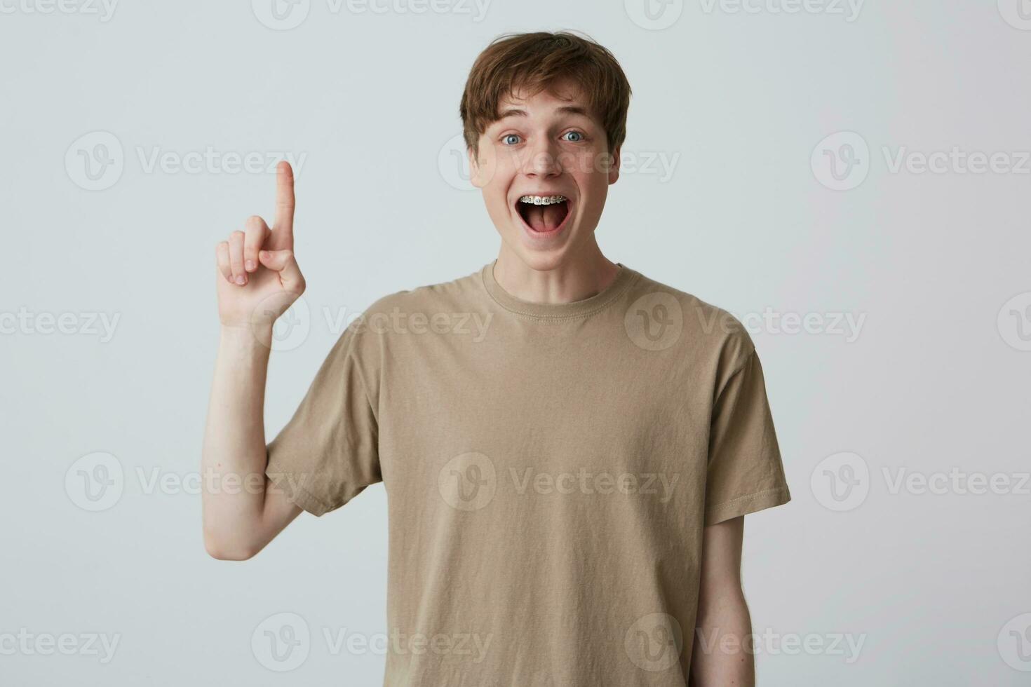 Happy excited blond young man with short haircut and braces on teeth wears beige t shirt and pointing up isolated over white background Have a new idea photo