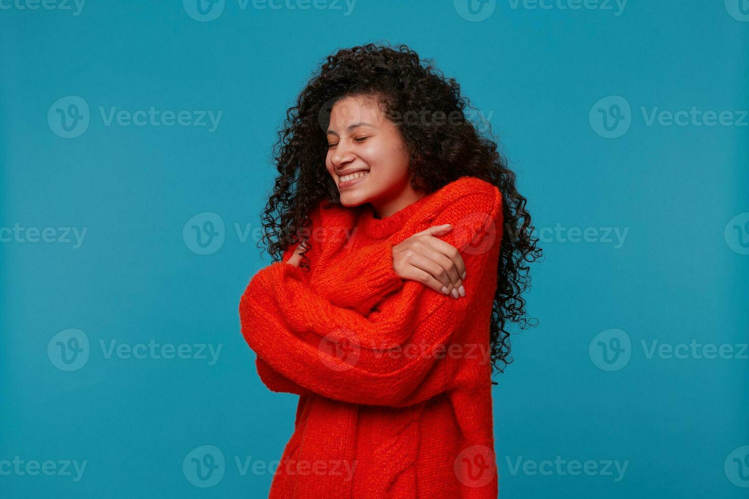 Fashion portrait of woman wearing warm oversized red knitted sweater, hugs herself tight with hands closed her eyes feels happy, over blue background photo