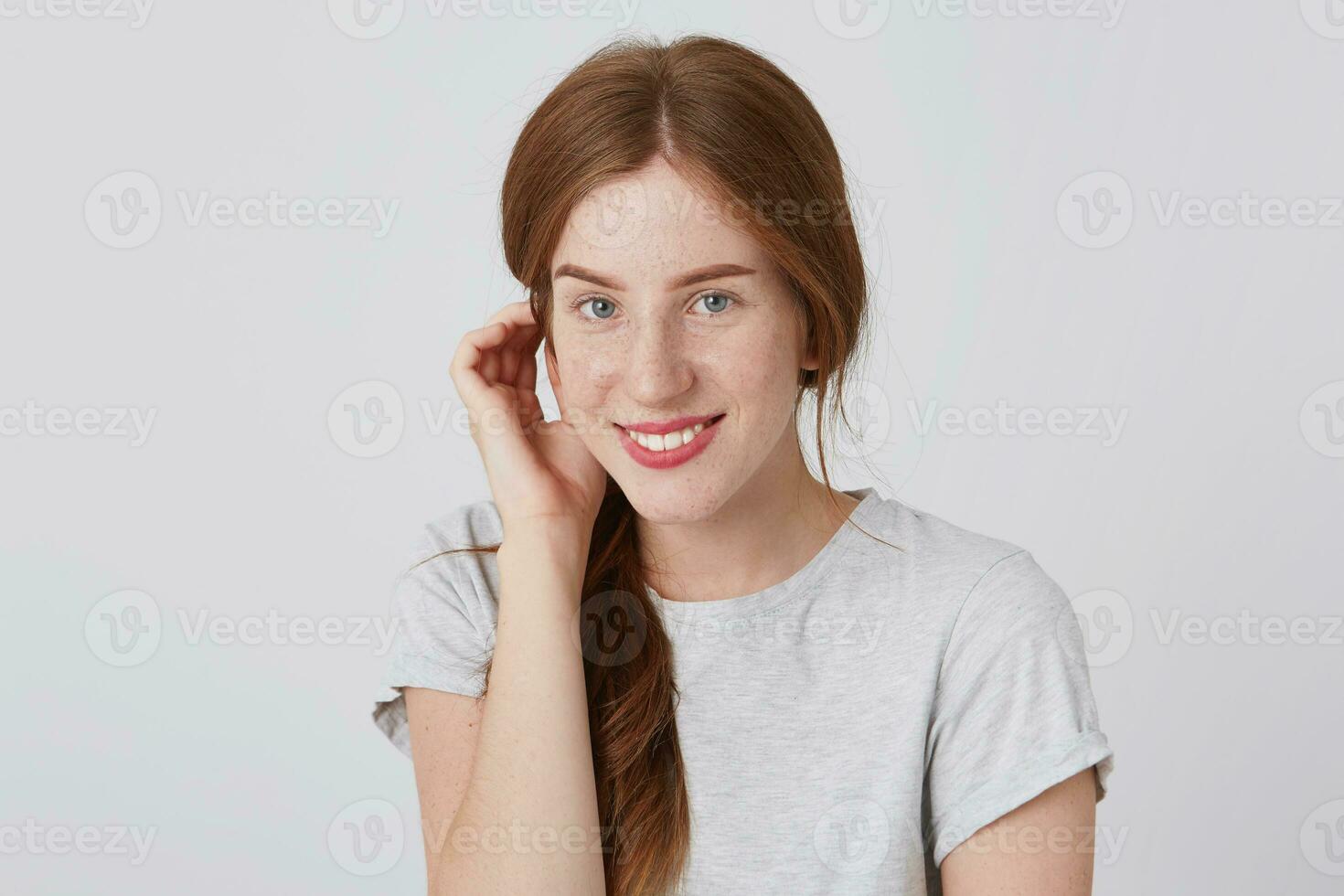 Portrait of happy lovely redhead young woman with freckles wears gray t shirt touching her ear and looks directly in camera isolated over white wall photo