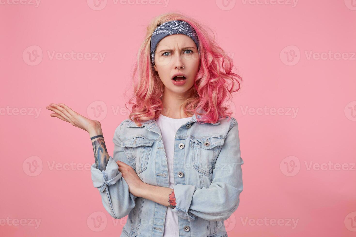 Young beautiful outraged pink haired woman in denim jacket, frown and indignant looking at the camera, wide open mouth in misunderstanding expression. Stands over pink background. photo