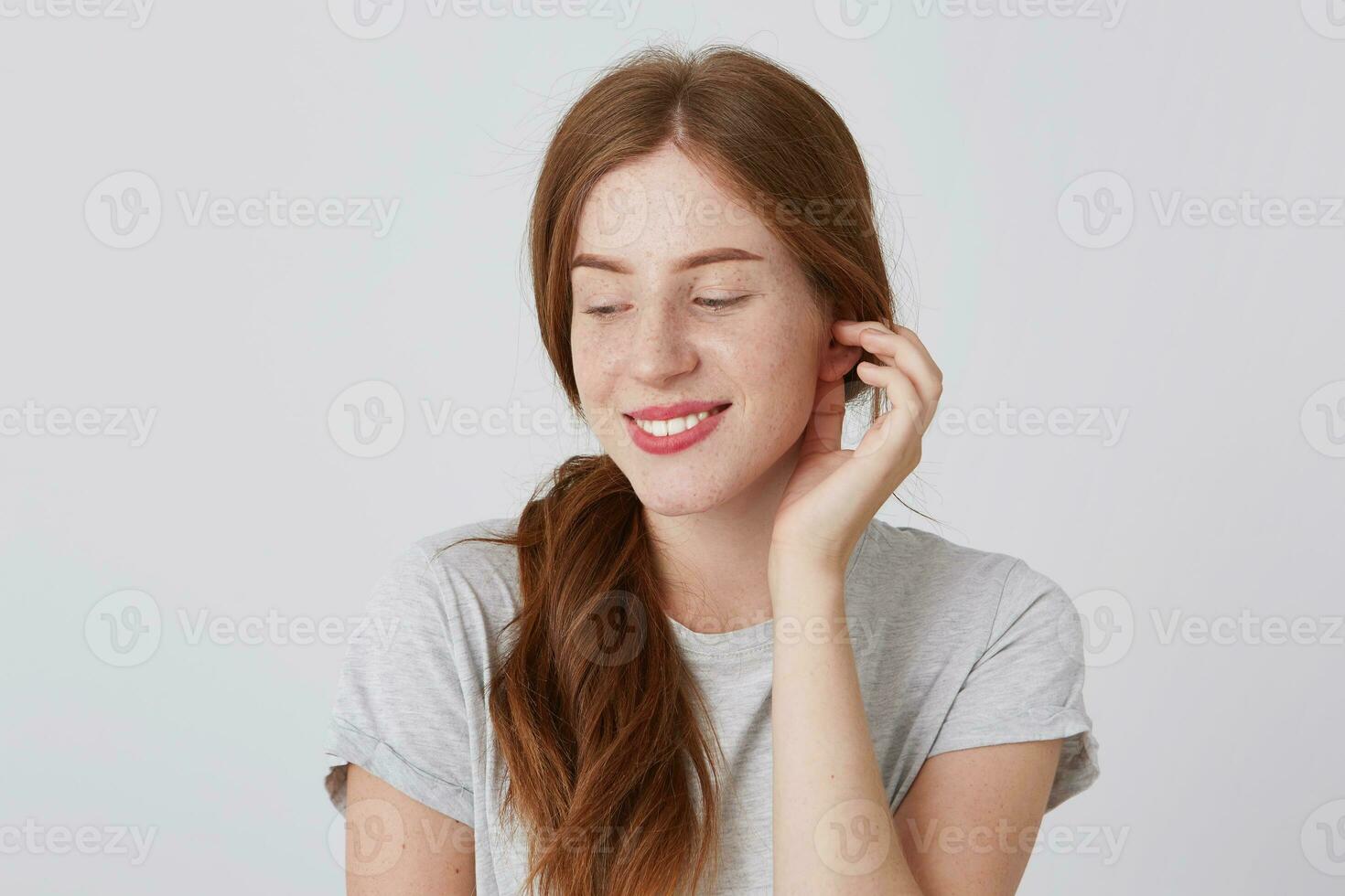 Cheerful timid young woman with red hair and freckles wears gray t shirt smiling and looking down isolated over white background Touching her ear photo