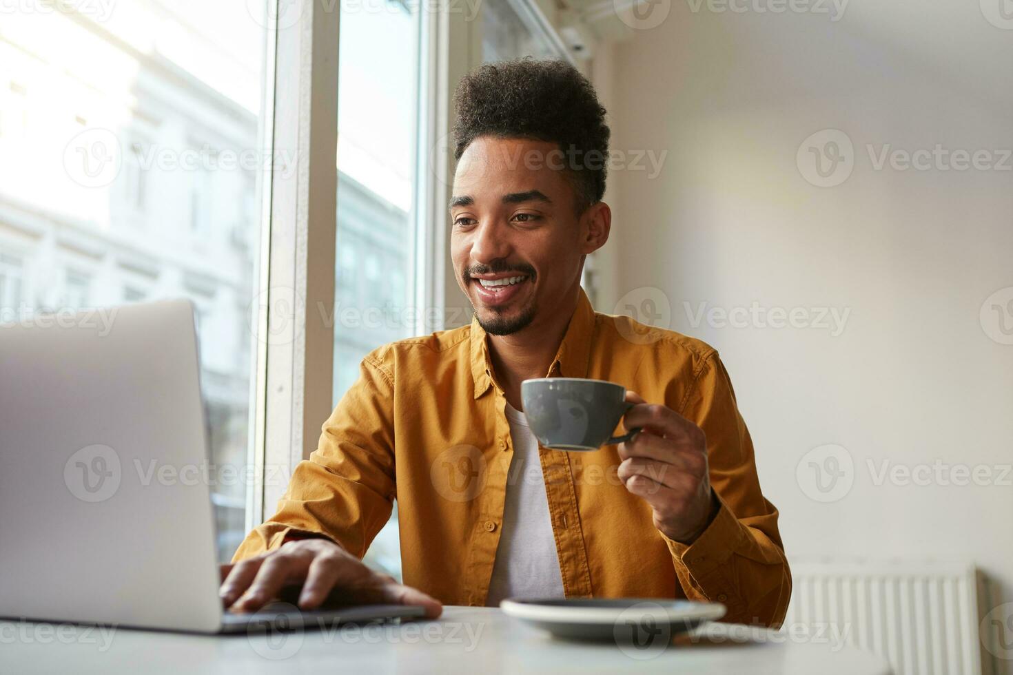 Young dark skinned guy sitting in a cafe and working on a laptop, wears in yellow shirt, drinks aromatic coffee , broadly smiling and enjoys the work of a freelancer. photo