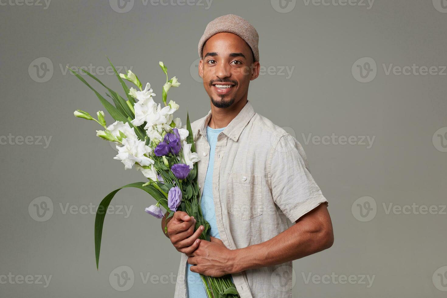 Young cheerful attractive man in gray hat, holds a bouquet in his and posing, looks at the camera with glad expression and smiling, stands over gray backgroud. photo