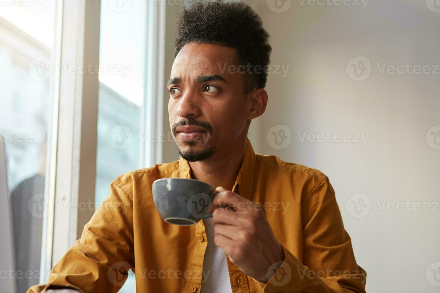 joven africano americano hombre usa en amarillo camisa, sentado a un mesa en un café y bebidas aromático café, pensando acerca de dónde a Vamos esta fin de semana. mirando pensativamente dentro el distancia. foto
